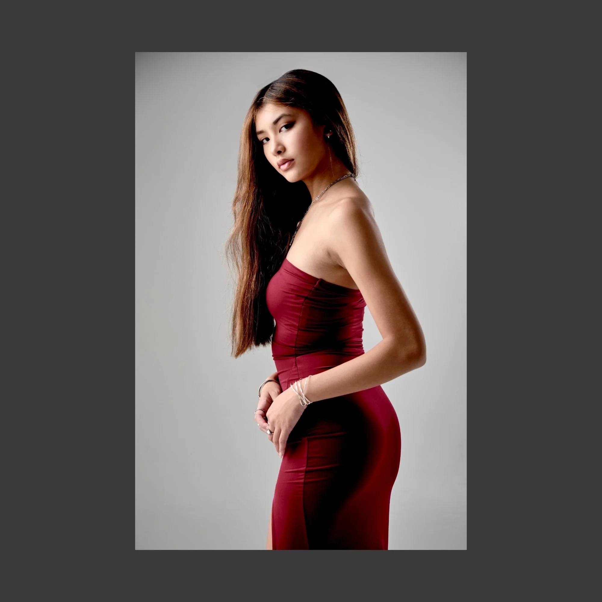 A young woman with long dark hair wearing a strapless, form-fitting red dress, standing against a plain gray background, looking at the camera with a neutral expression.