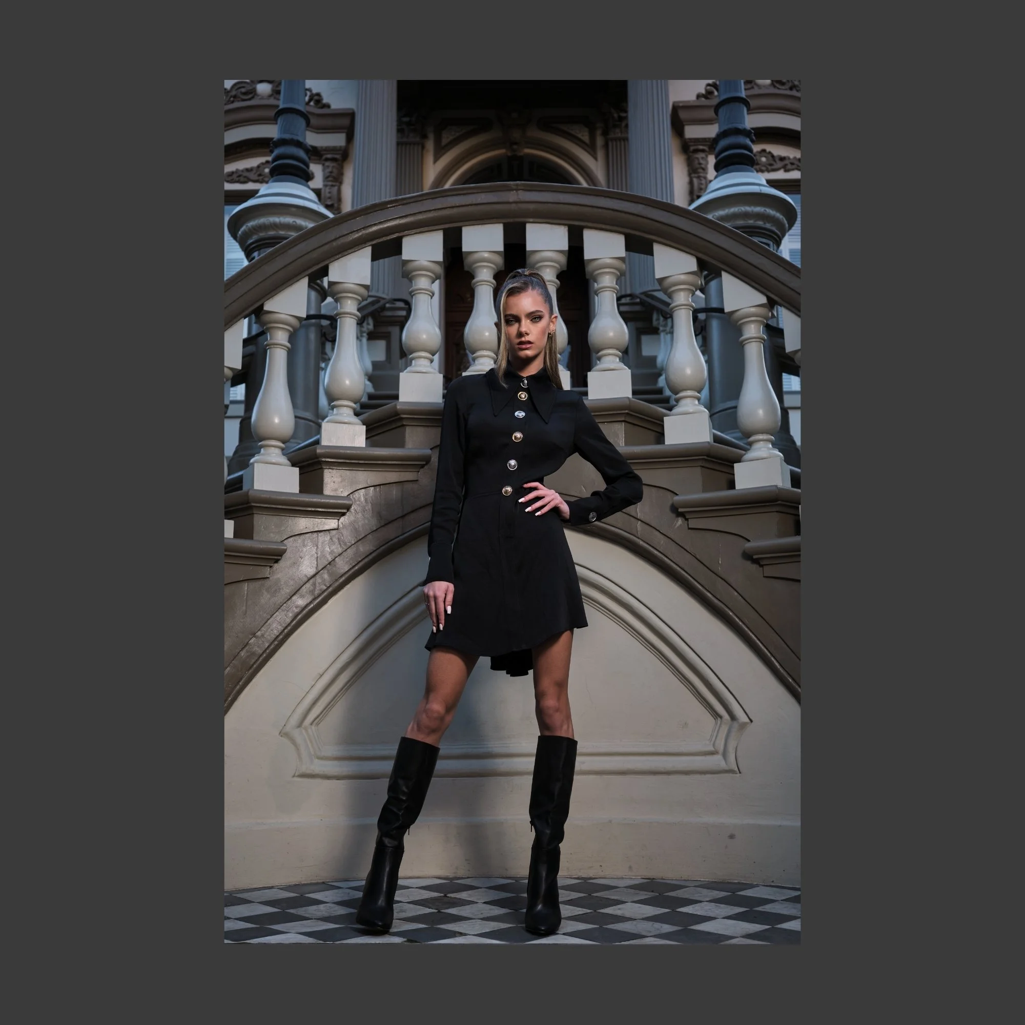 A woman in a black dress with gold buttons, wearing black high-heeled boots, stands confidently on a checkerboard floor in front of an ornate staircase and historic building.