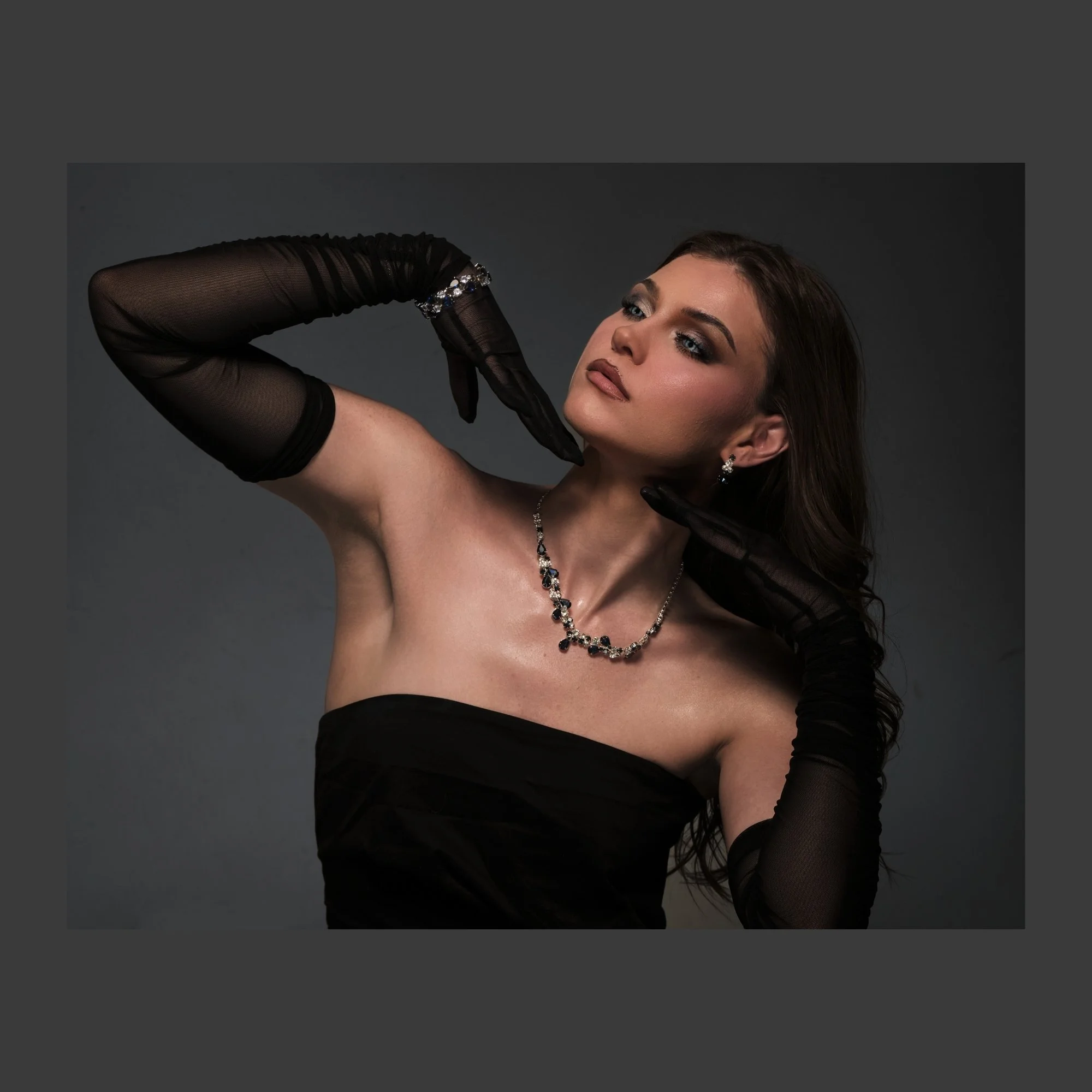 A woman with long brown hair and makeup wearing a black strapless dress, black gloves, and jewelry, posing against a gray background.
