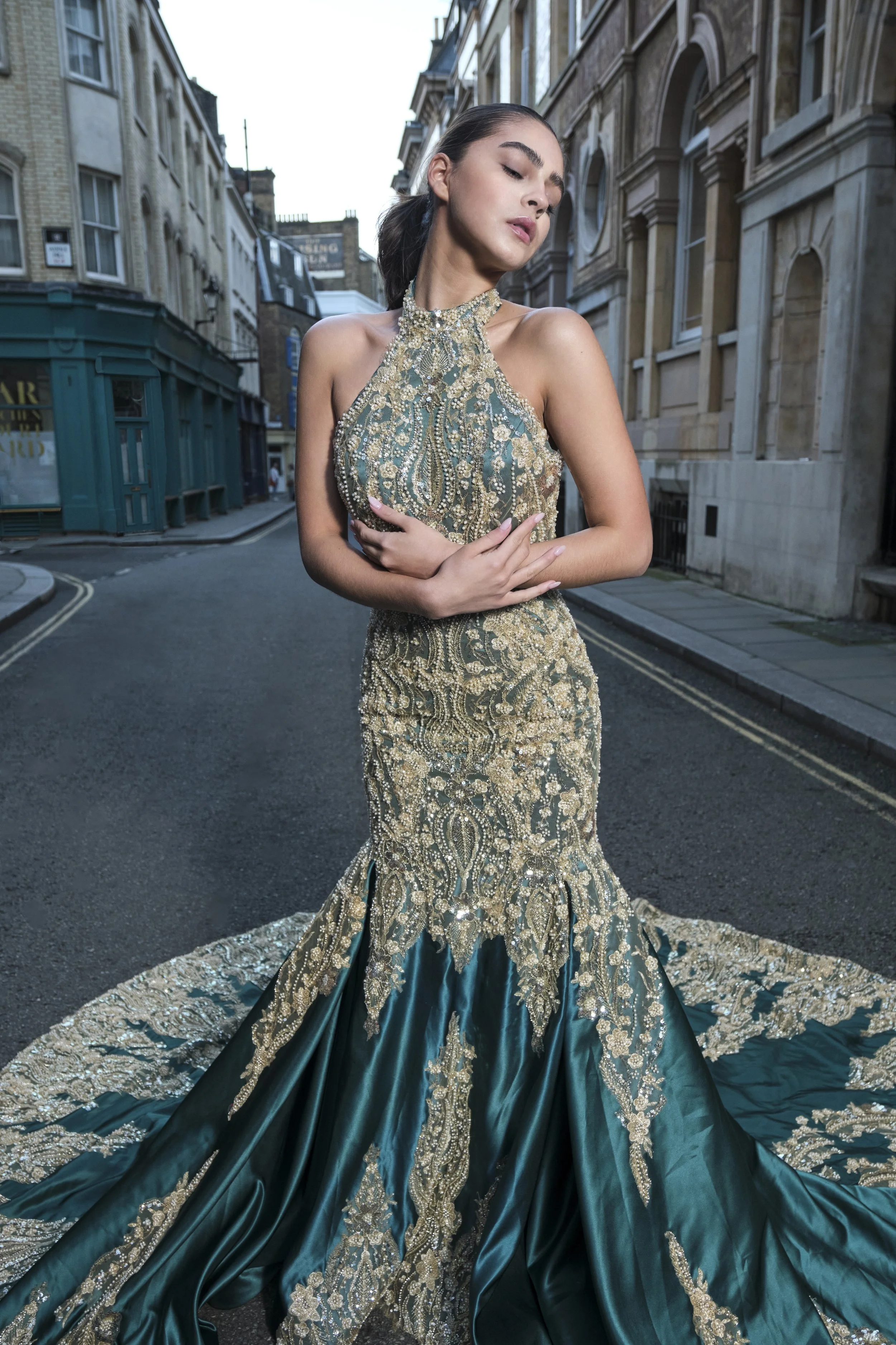 A woman in a luxurious, embroidered gown standing on an empty city street, with her eyes closed and arms crossed over her chest.