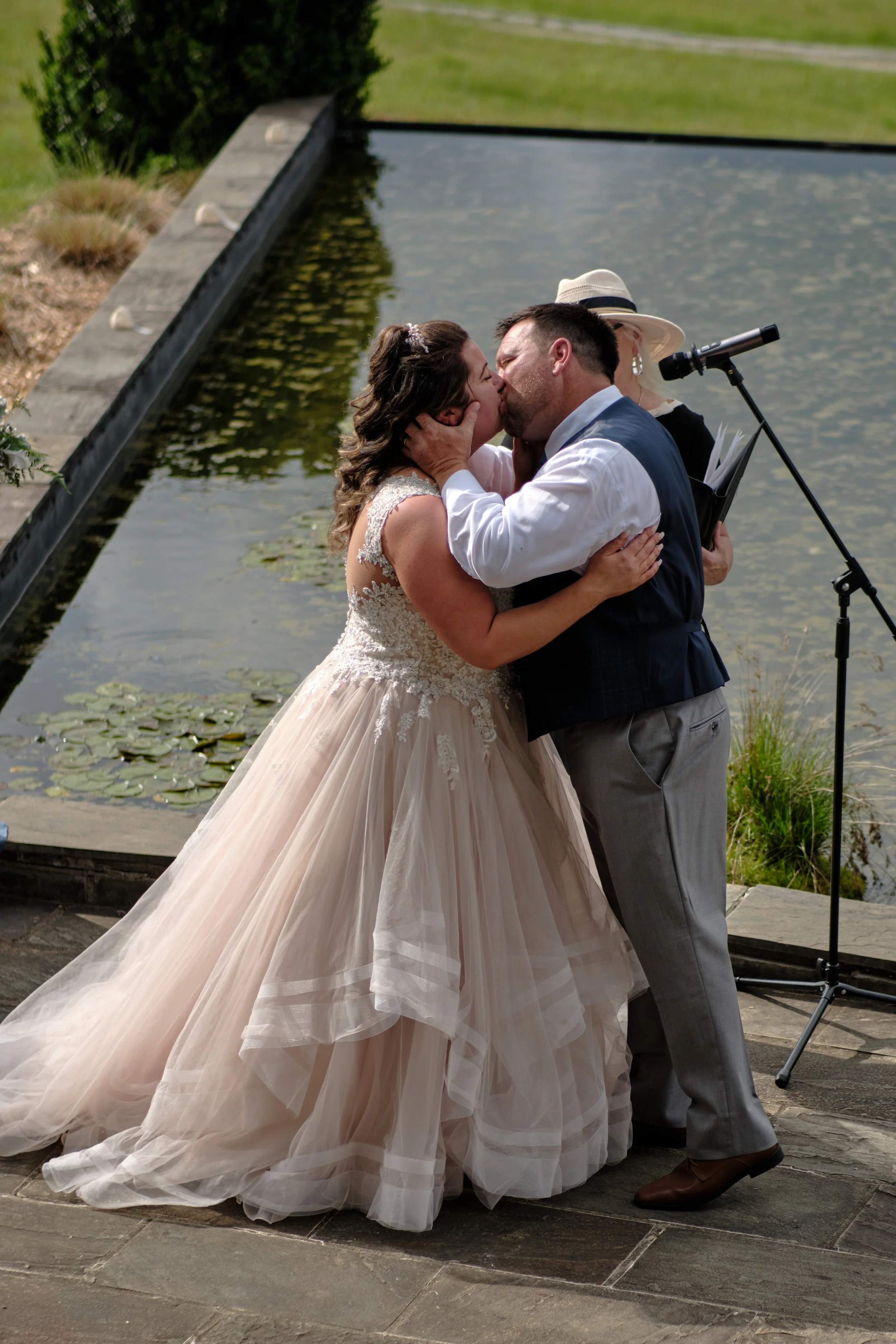 A bride and groom kissing during their wedding ceremony by a pond with water lilies.