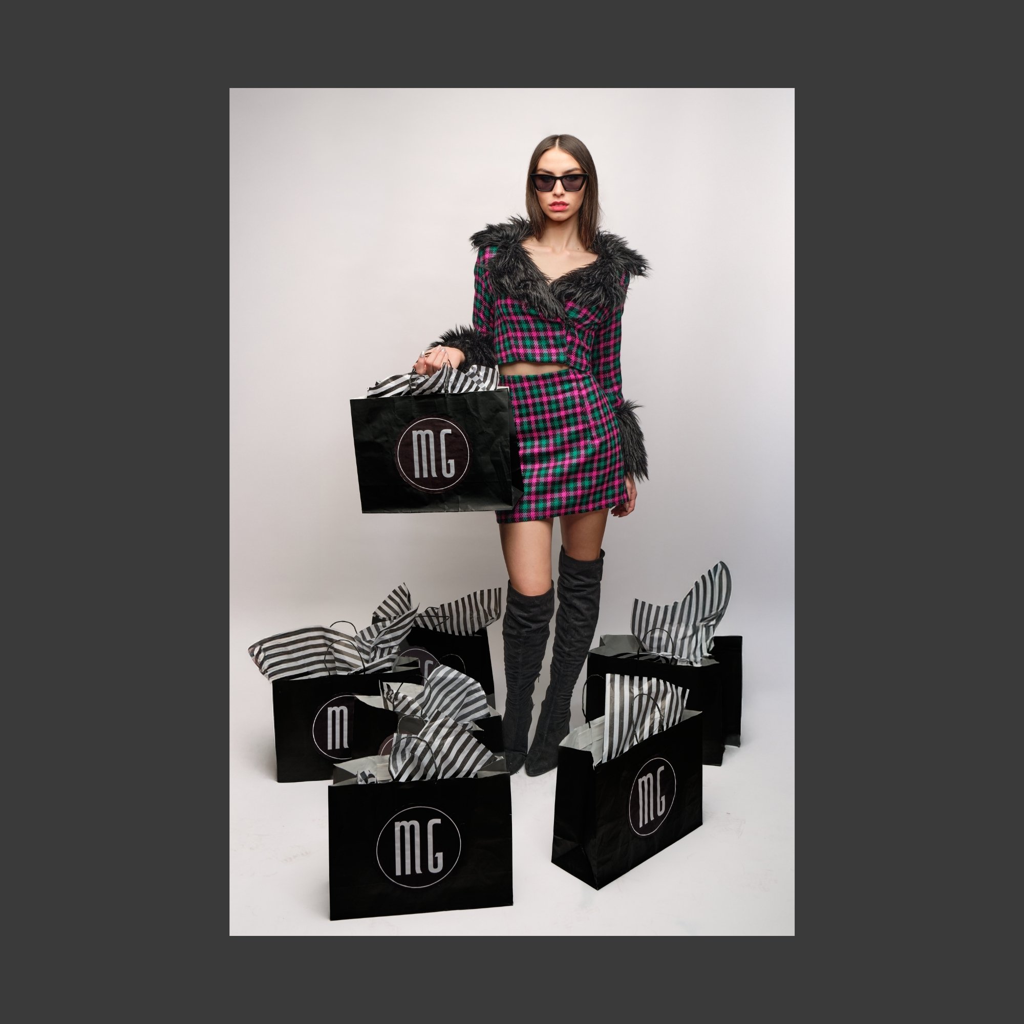 A woman wearing a pink and green plaid outfit with faux fur trim, black thigh-high boots, and sunglasses, holding a black shopping bag with a circular logo, surrounded by additional shopping bags with striped tissue paper.