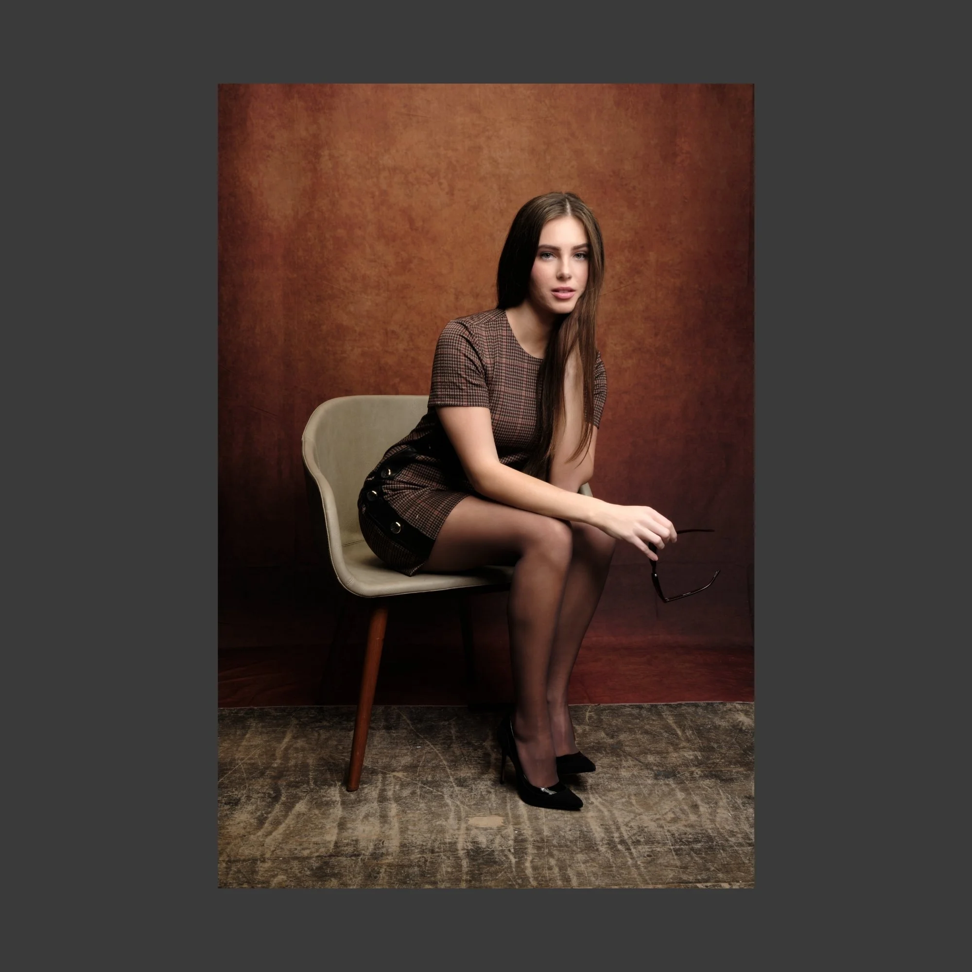A young woman with long brown hair, wearing a brown checkered dress, black stockings, and black high heels, sitting on a beige chair against a brown textured background, holding sunglasses in her right hand.