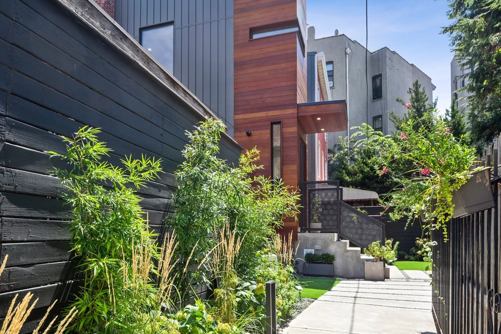Modern townhouse with wooden exterior and black fence, surrounded by lush green plants and flowers, sidewalk leading to stairs, outdoor greenery and trees.