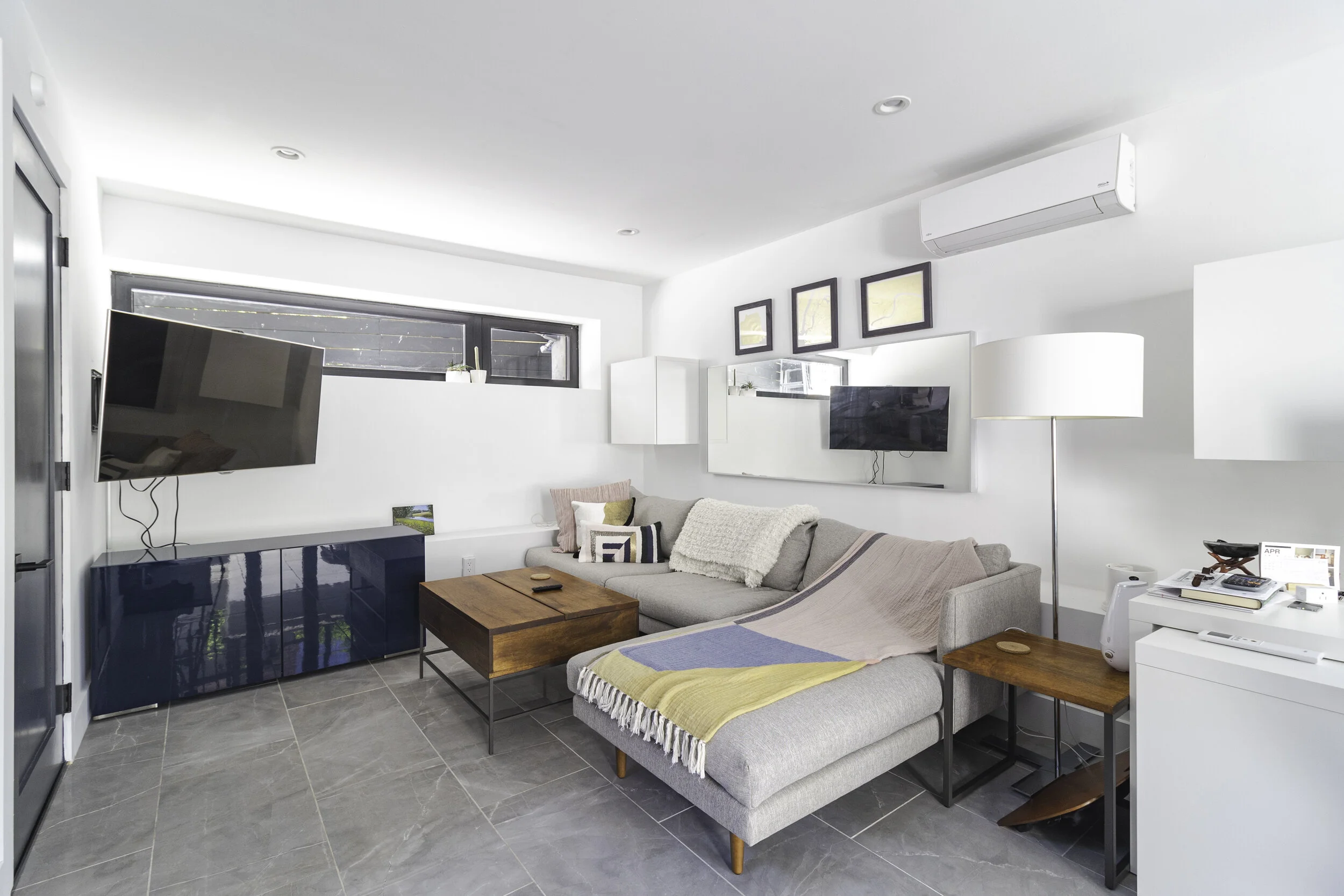 Modern living room with sectional gray sofa, wooden coffee table, large wall-mounted TV, smaller TV above sofa, white walls, tiled floor, and window with black blinds.
