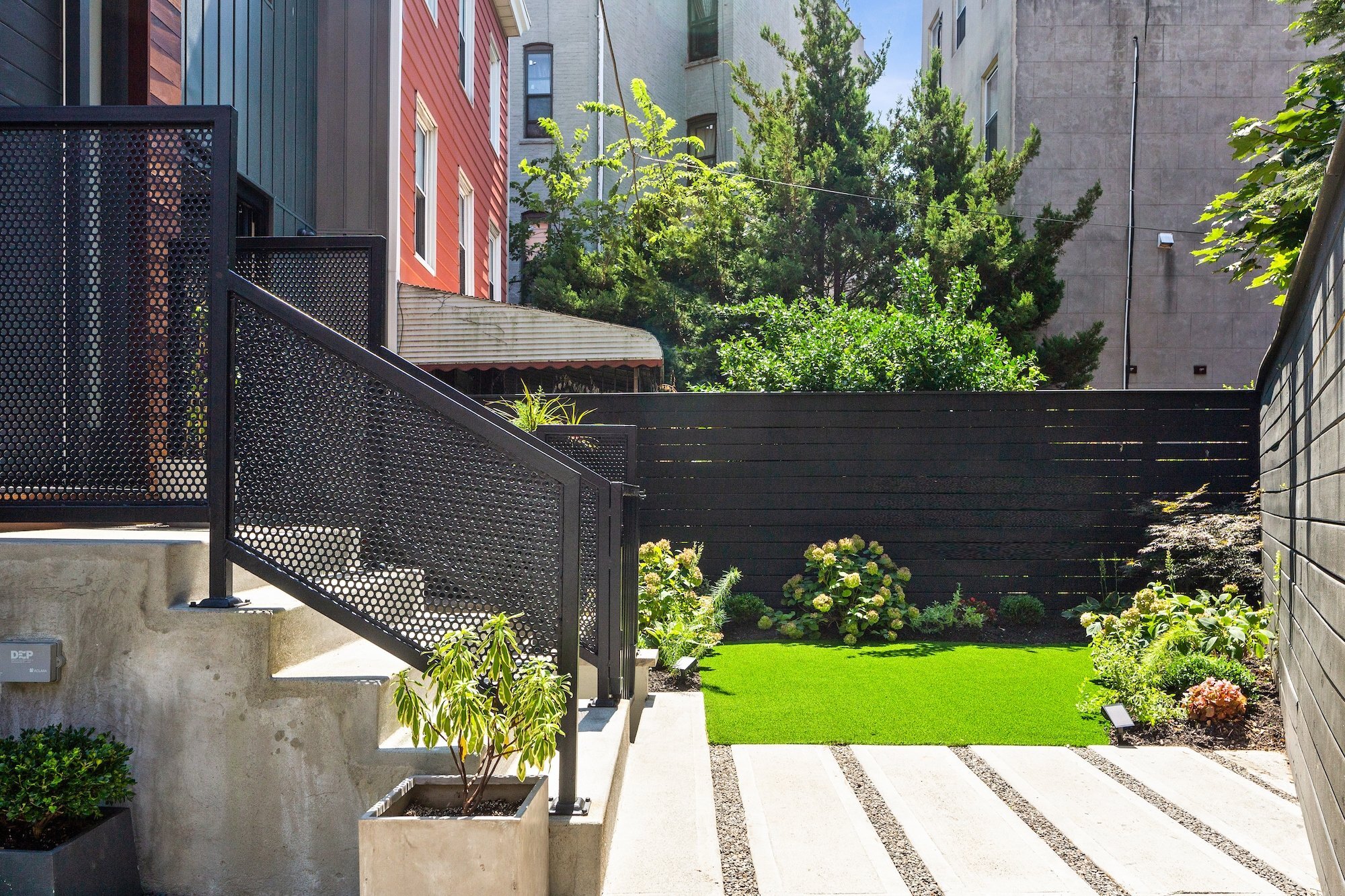 Urban backyard with modern design, black metal staircase, green lawn, potted plants, and surrounding buildings