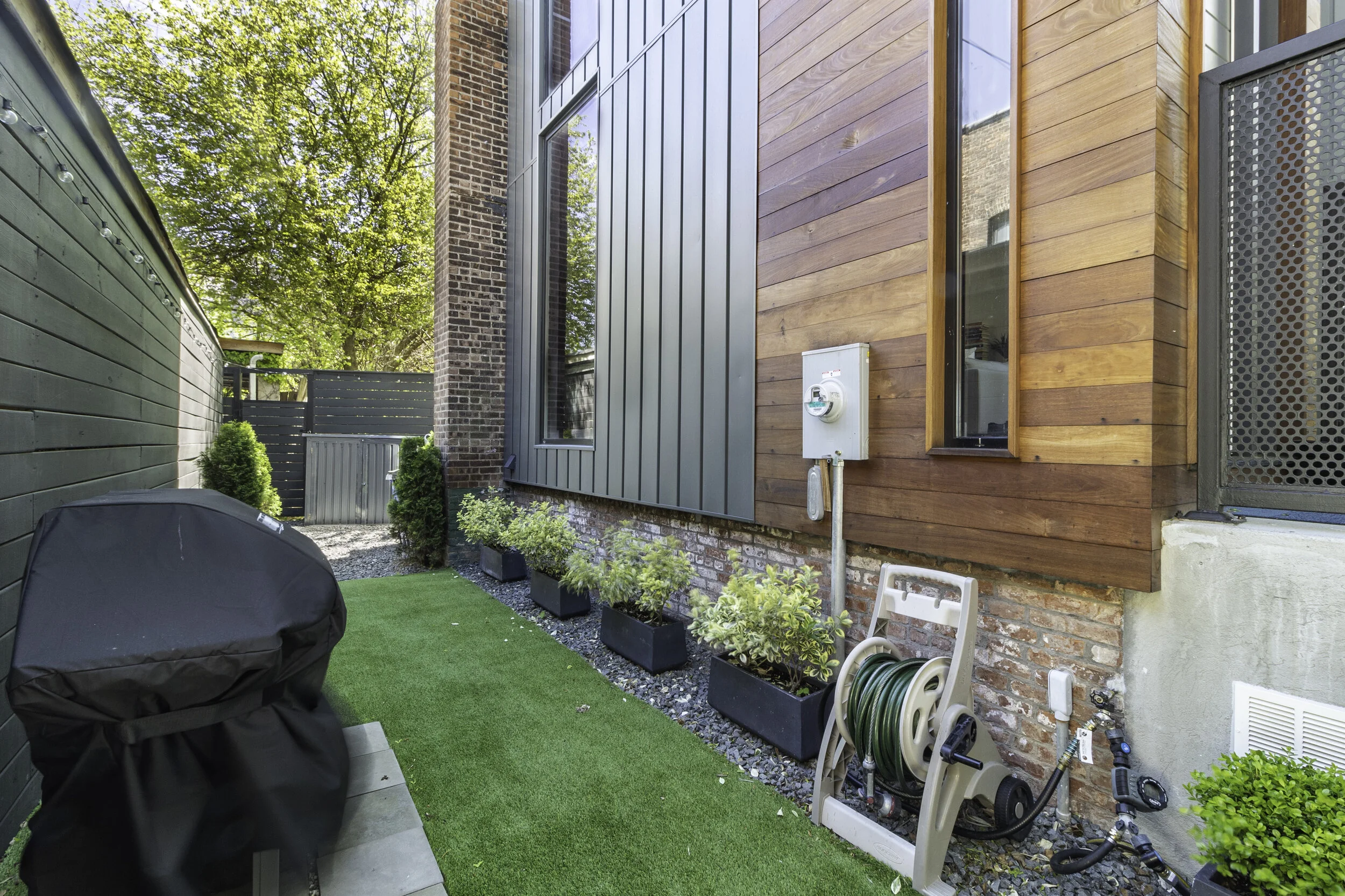 Side yard with artificial grass, plants in rectangular pots, barbecue covered with a black cover, water hose reel, electrical box mounted on the house, windows, and trees in the background.