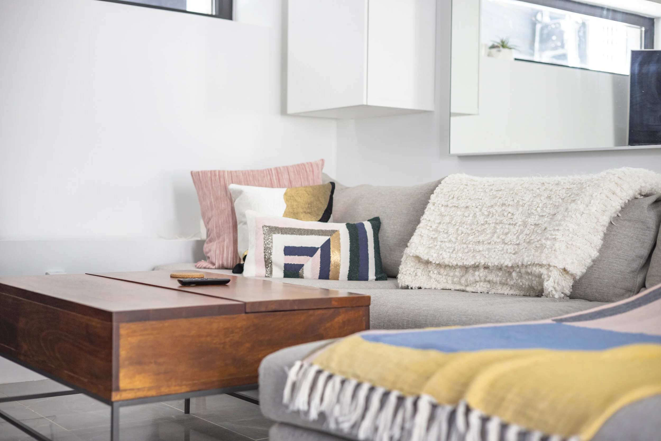 Living room with gray sofa decorated with colorful and textured pillows, a wooden coffee table, and a white blanket. There is a large mirror on the wall and a window with a small plant visible outside.