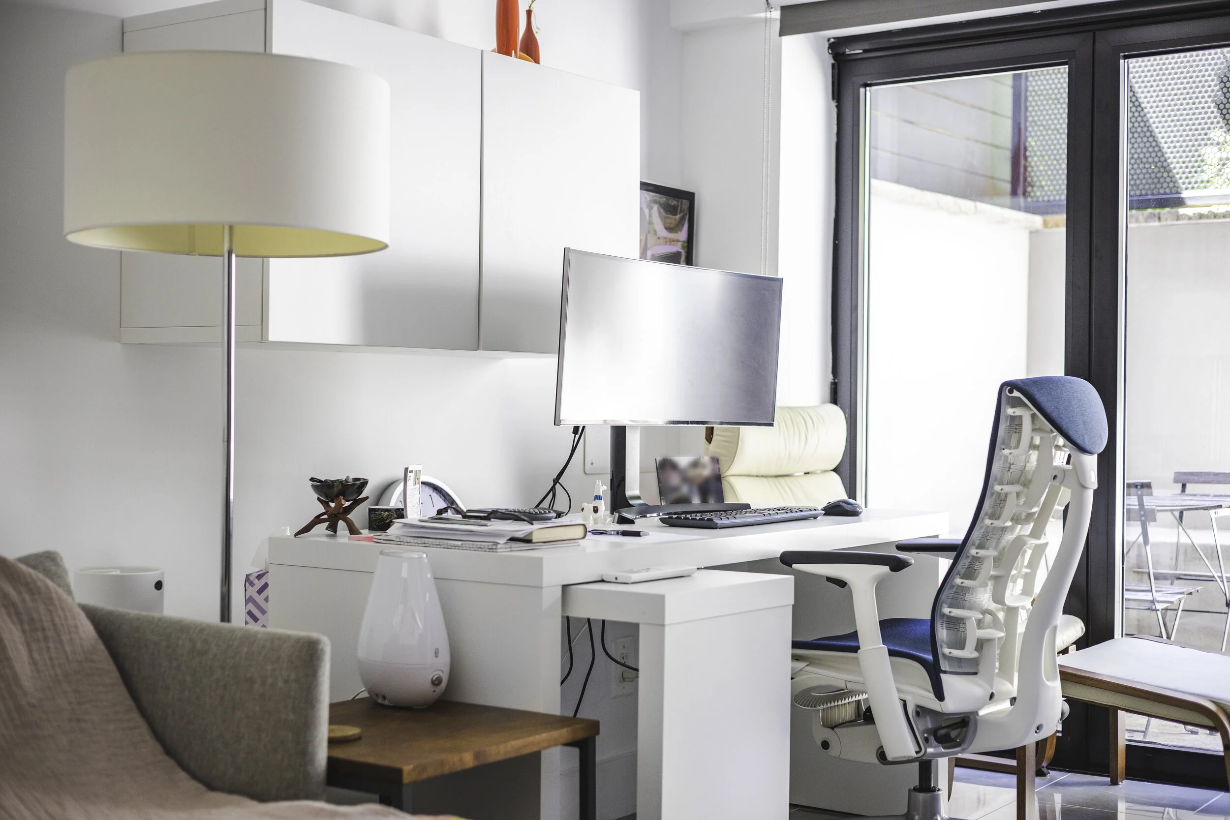 Modern home office with a white desk and ergonomic chair, computer monitor, and large window with sliding glass door