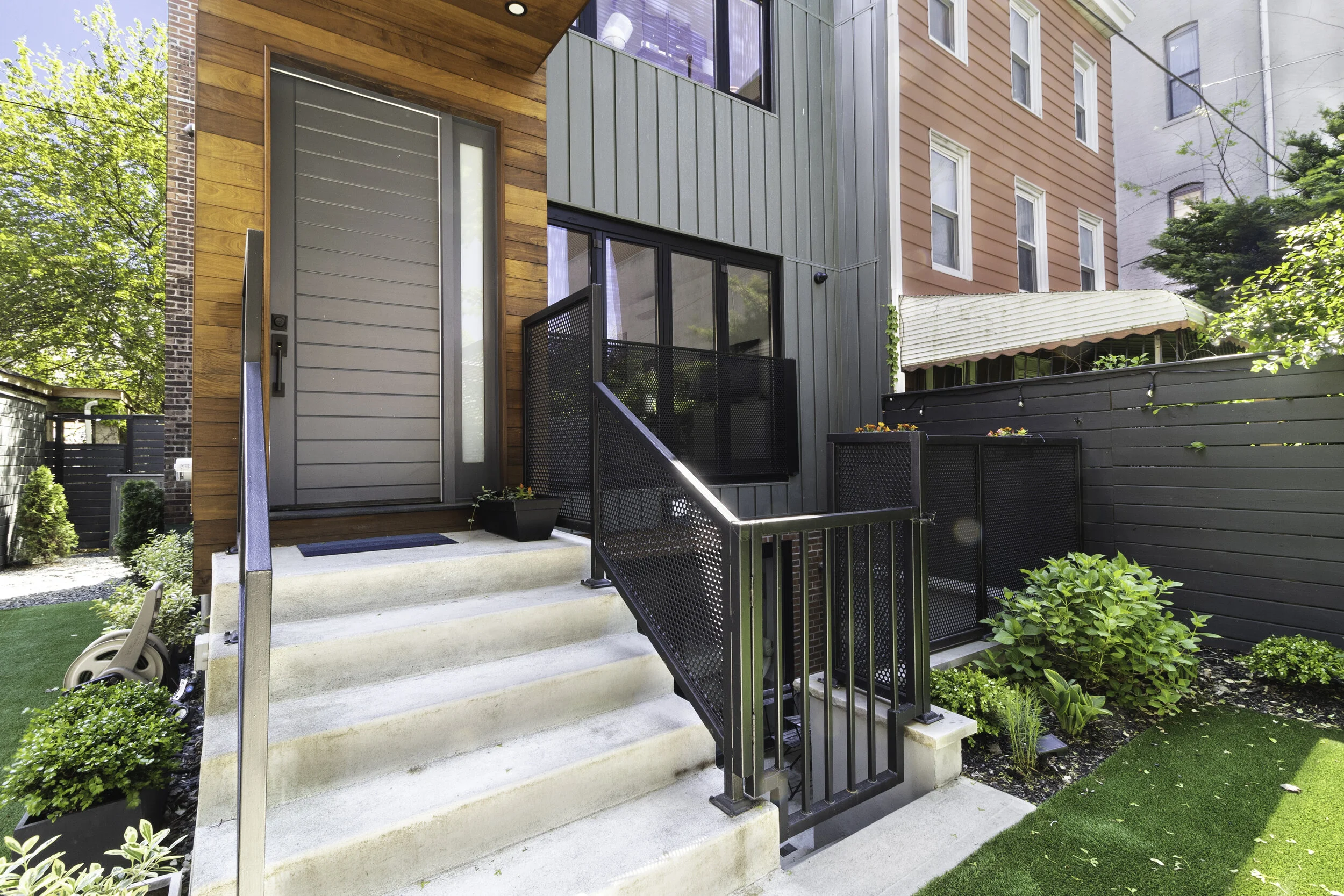 Townhouse entrance with concrete steps, gray door, black metal railings, greenery, and a modern exterior with ipe wood and zinc siding.