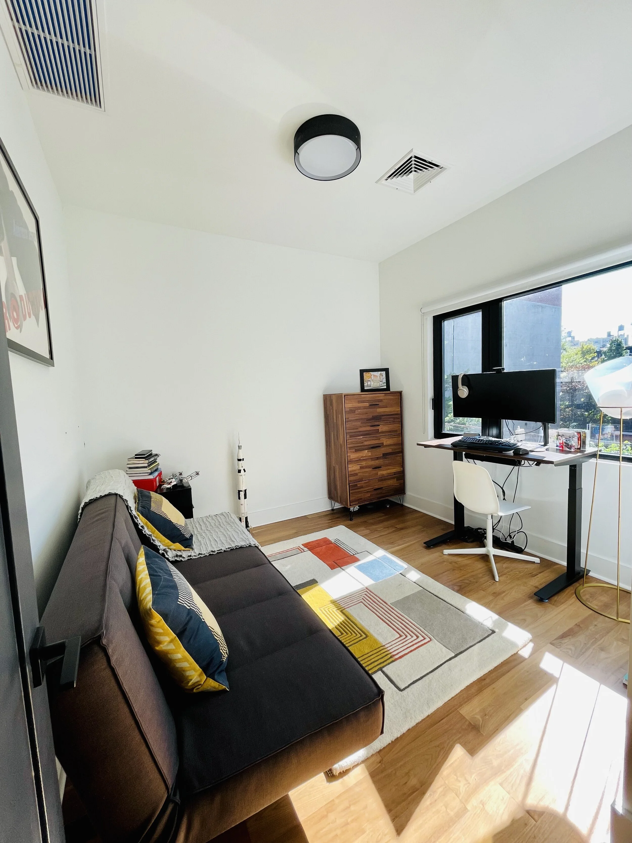 A bright living room with a dark sofa, colorful pillows, a patterned rug, a desk with a large monitor, a white chair, a wooden cabinet, and a window with a city view.