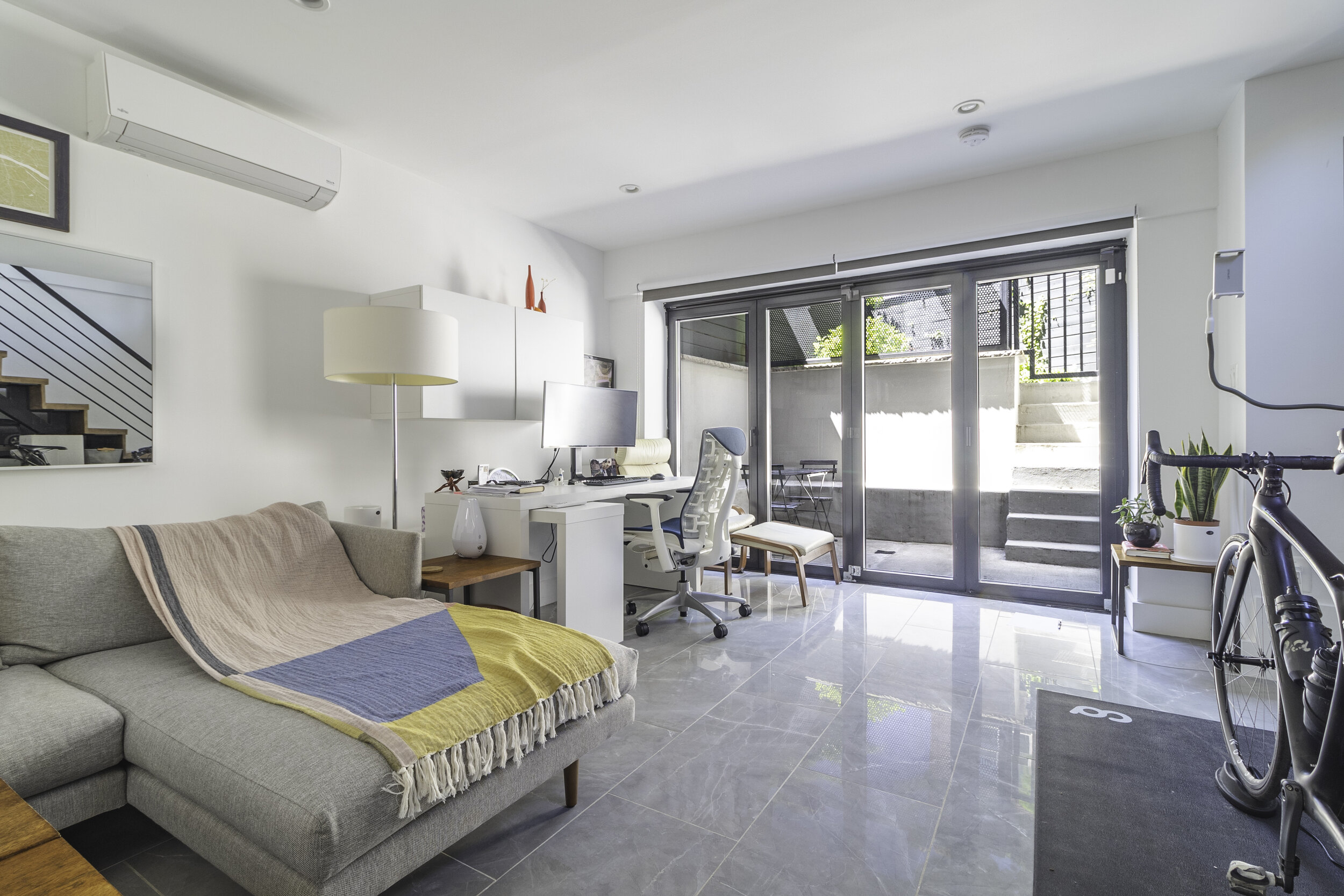 Bright living room with gray sofa, beige and blue blanket, white desk with computer, sliding glass door to outdoor patio, indoor plants, bicycle, and staircase reflected in a mirror.