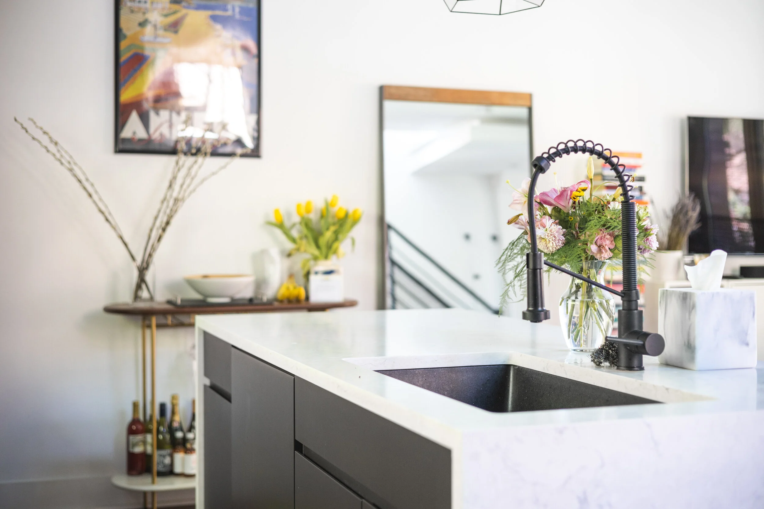 Modern kitchen with white marble countertop, black sink, black faucet, and floral arrangements in vases. In the background, there is a wooden console with yellow tulips and a framed abstract art piece on the wall.