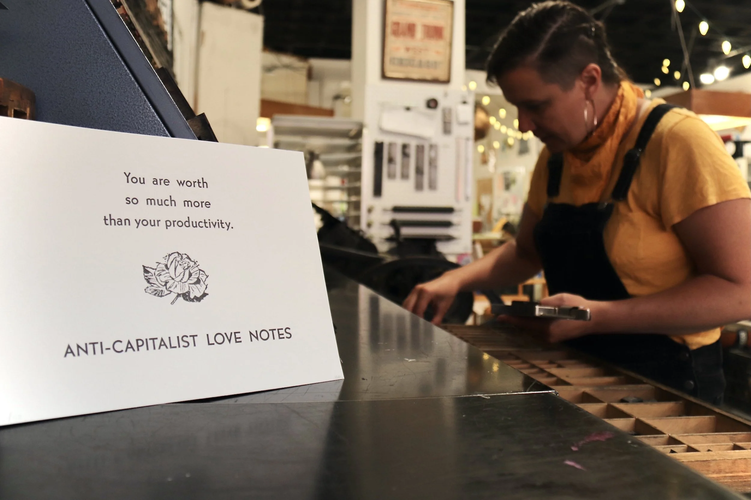 nicole setting type at the printshop, with a print in the foreground that says, "you are worth so much more than your productivity. anti-capitalist love notes."