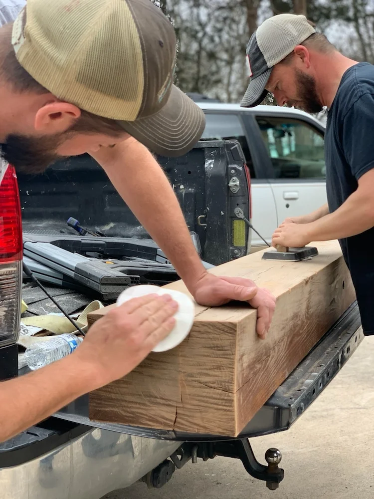 Two men working on a large piece of wood in the back of a pickup truck, with tools and a water bottle nearby, outdoors with trees in the background.