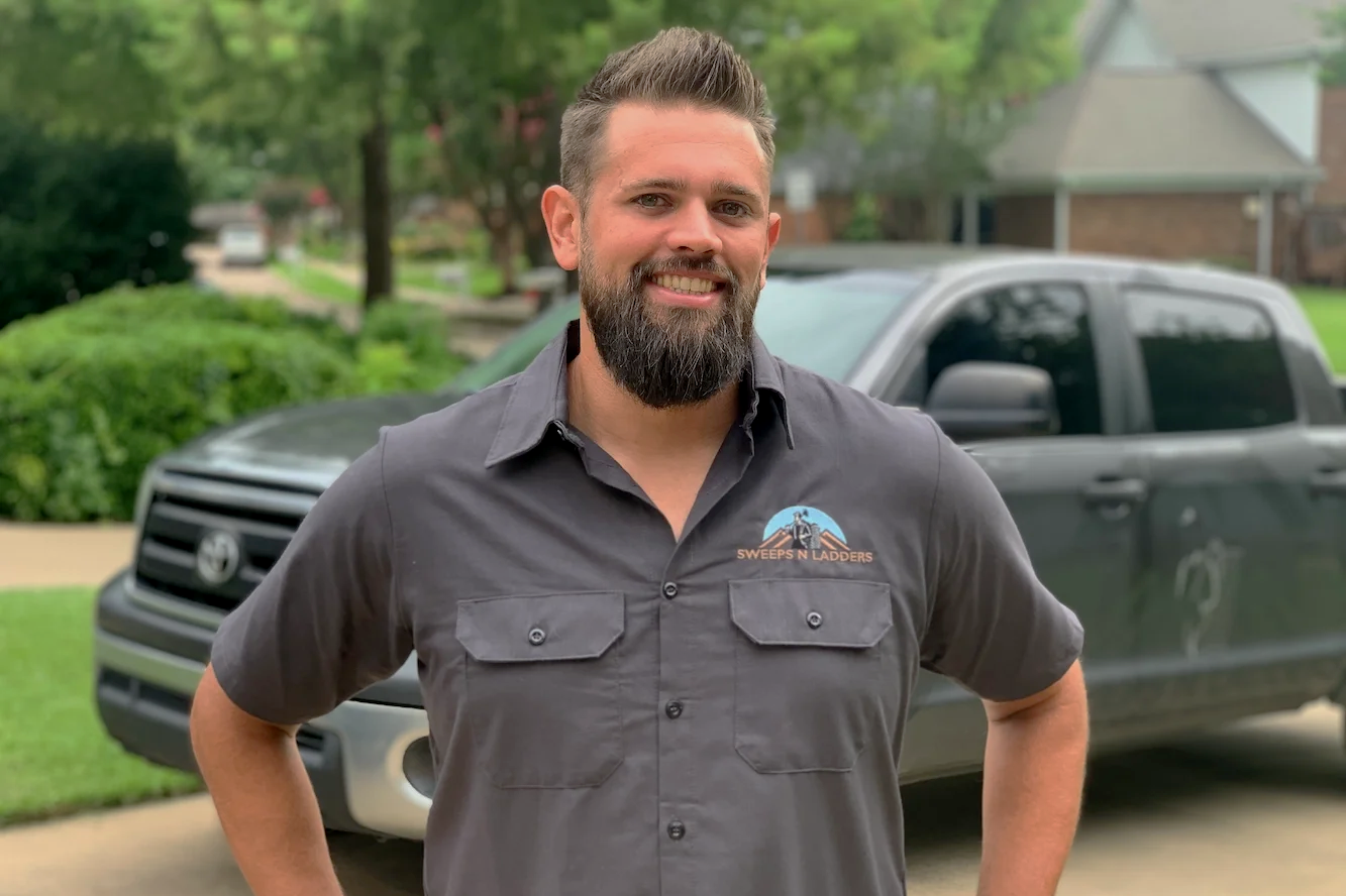 Man with beard smiling, wearing a gray shirt with 'Sweeps N Ladders' logo, standing outdoors in front of a black pickup truck.