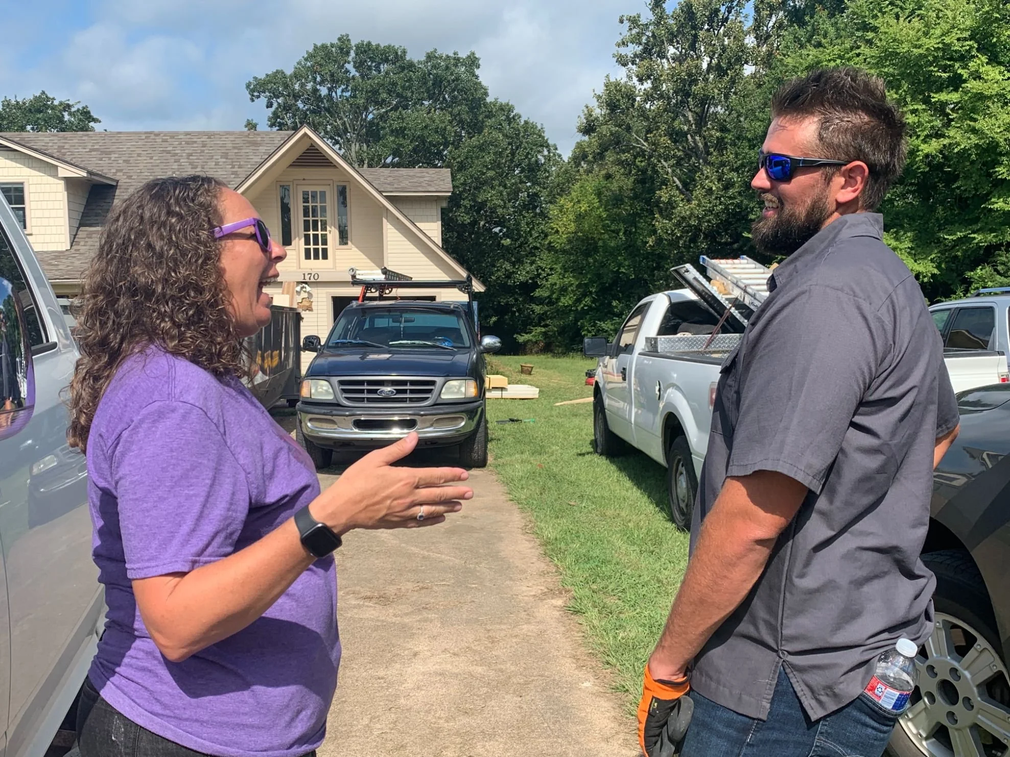 Two people, a woman with curly hair in a purple shirt and a man with a beard in a gray shirt, are having a conversation outdoors in a driveway. They are smiling and wearing sunglasses, with multiple vehicles and a house in the background.