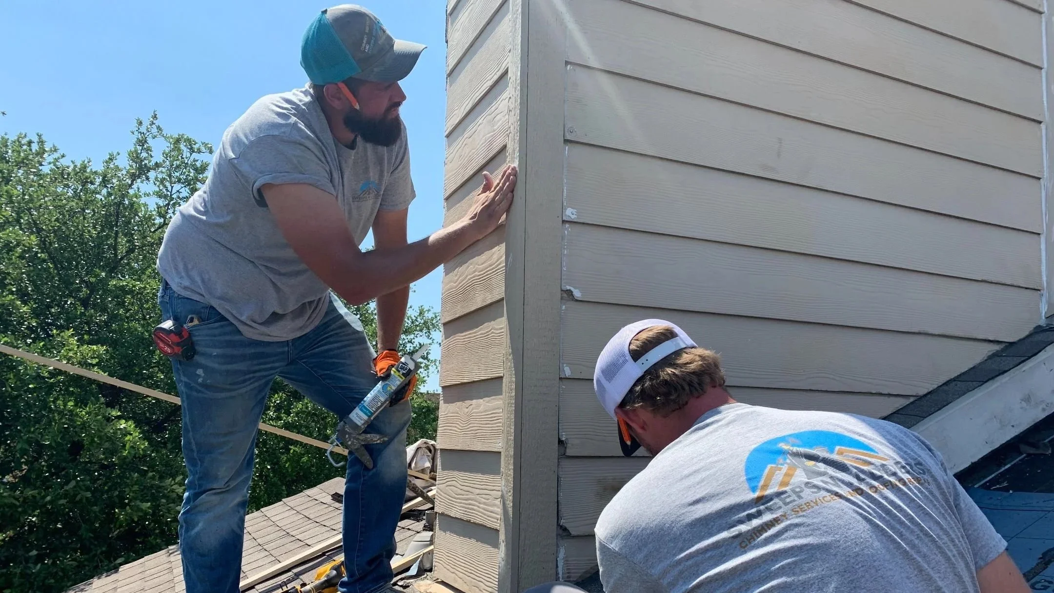 Three workers installing or repairing siding on a small rooftop structure on a beige house. One worker is on a ladder using a power drill, while the other two stand nearby.