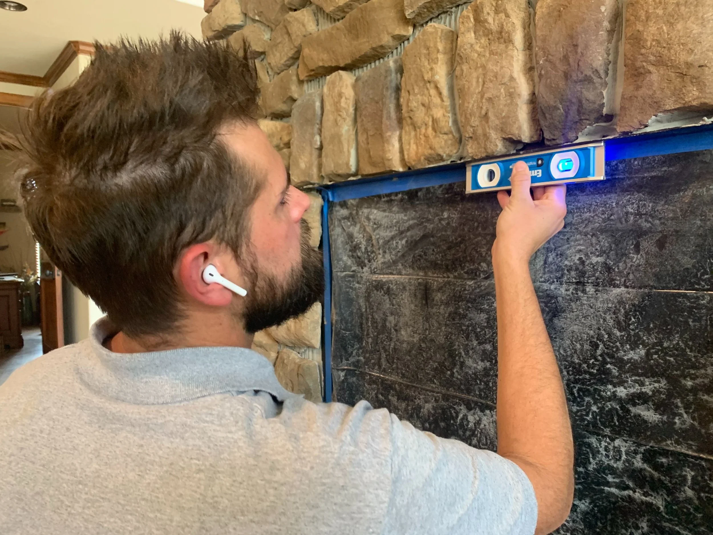 Man measuring a wall with a level tool, wearing wireless earbuds, wearing a light gray shirt, with a stone and ceramic tiled wall background.
