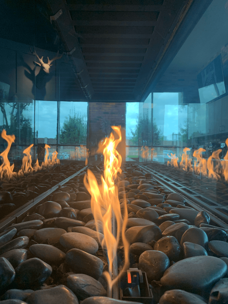 Indoor fireplace with glass walls, black rocks on the floor, visible flames, and a deer head mounted on the wall above.