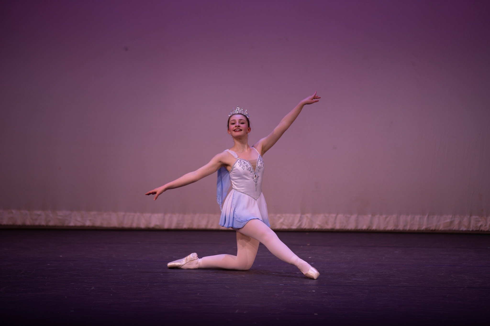 A young ballerina performing on stage, kneeling on one knee with arms extended gracefully, wearing a light blue tutu and a tiara.