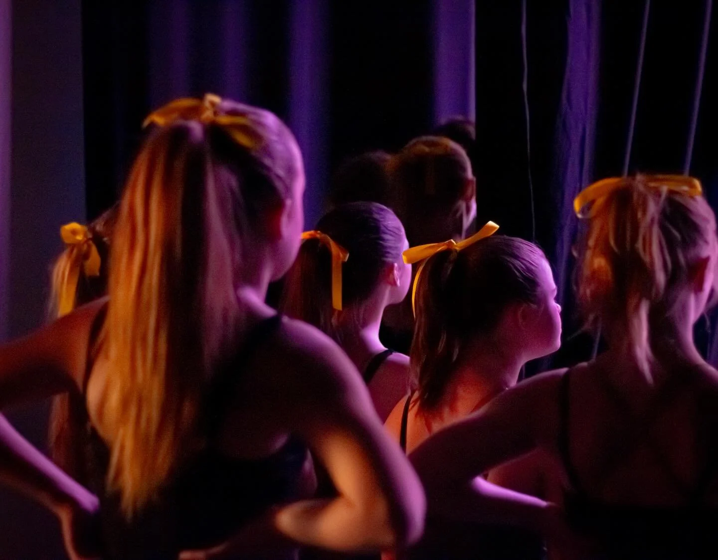 Group of young women seated backstage, with their hair tied with yellow ribbons, waiting for a performance, illuminated by purple stage lighting.