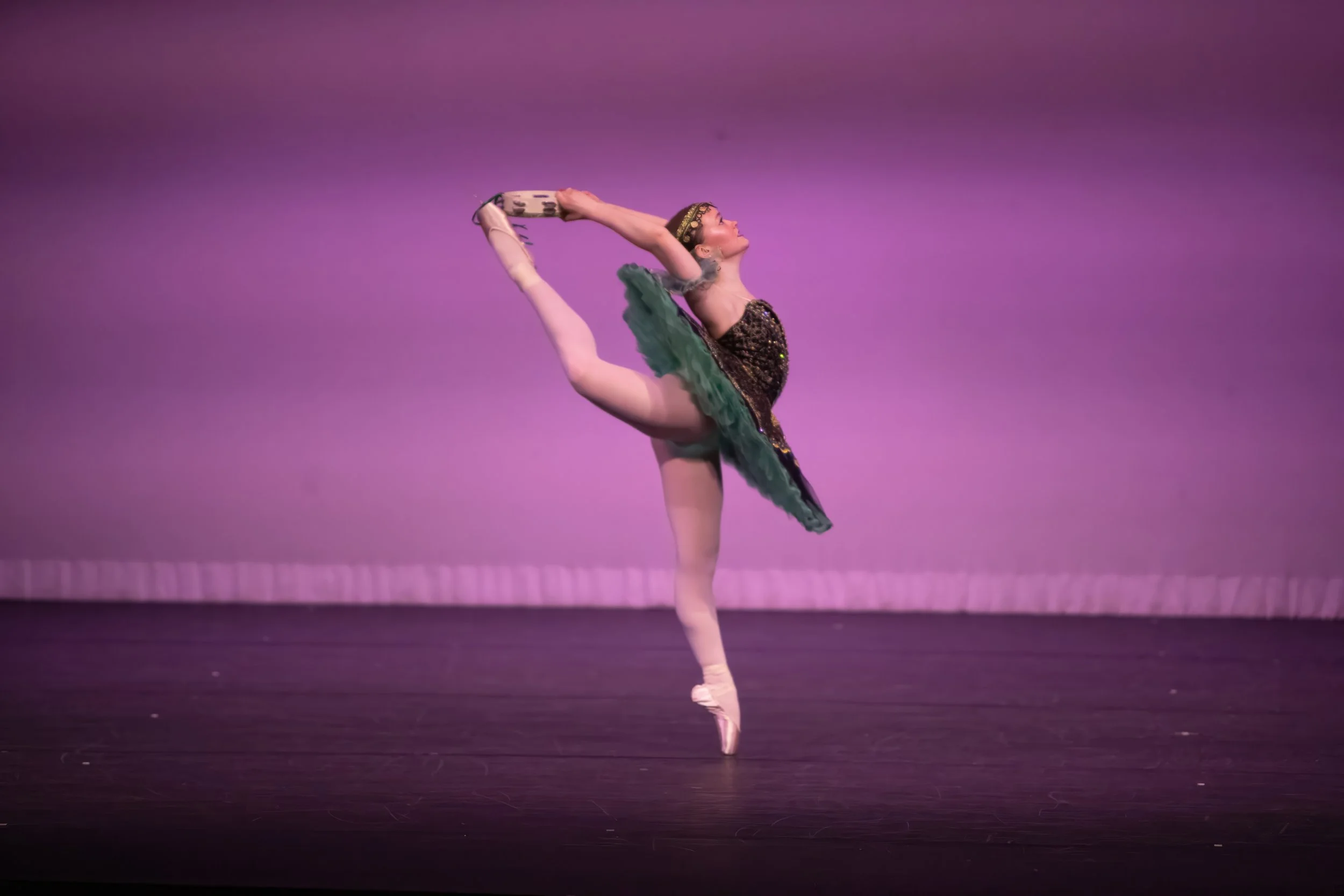 A ballet dancer in a costume performing a standing split on stage with a purple background.