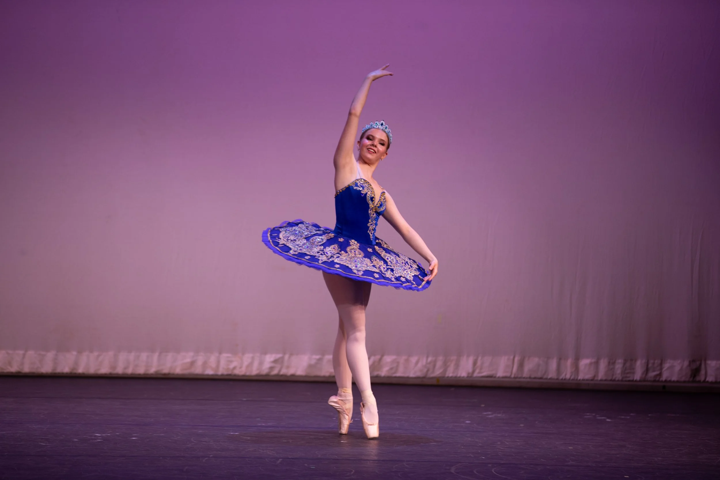 A ballerina performs on stage wearing a blue and gold tutu and a tiara, balancing en pointe with one arm raised.