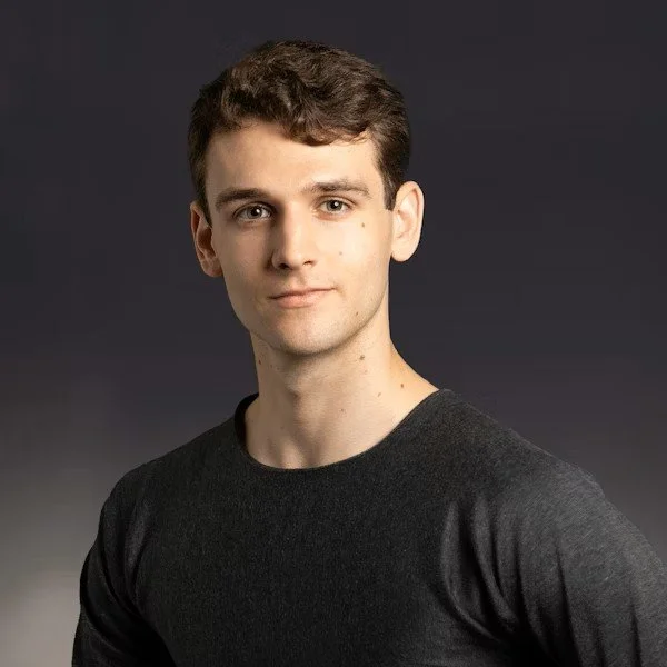 A man with short brown hair wearing a dark gray shirt, posing against a neutral gray background.
