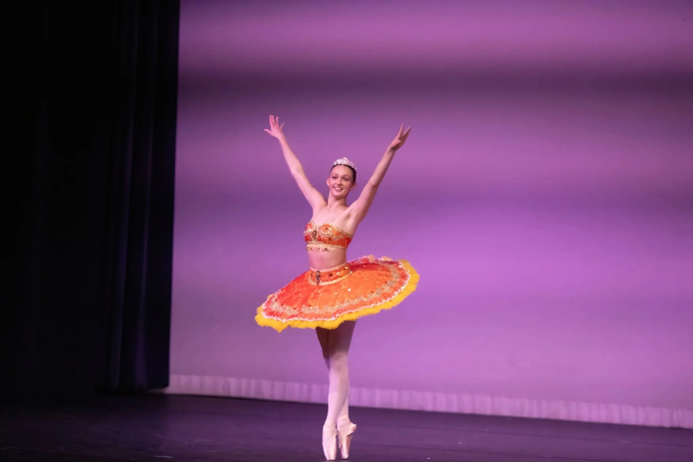Ballet dancer in a colorful orange tutu and tiara dancing on stage with a purple background.