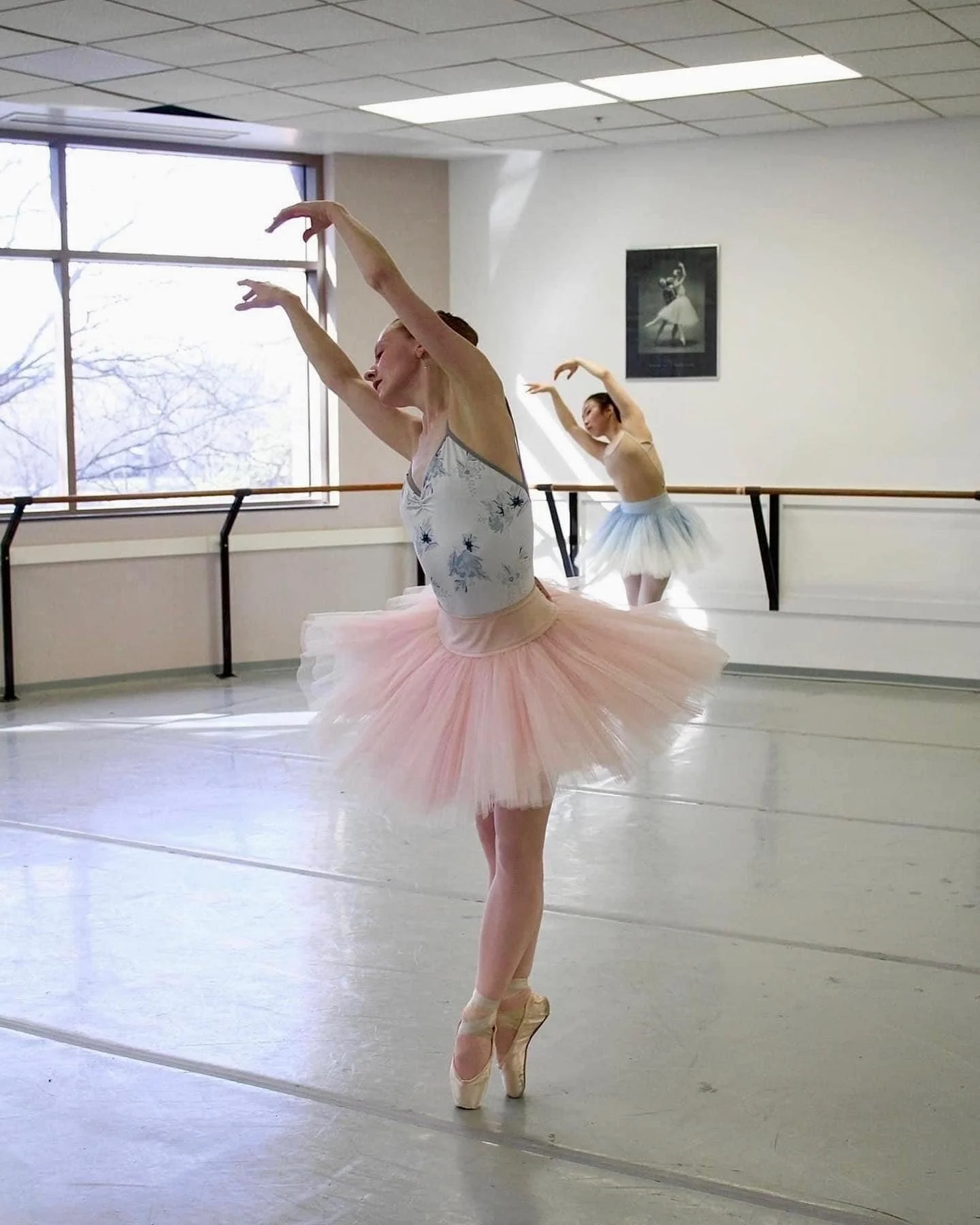 Ballet dancers practicing in a studio, one in pink tutu, another in blue, both on pointe with arms raised.