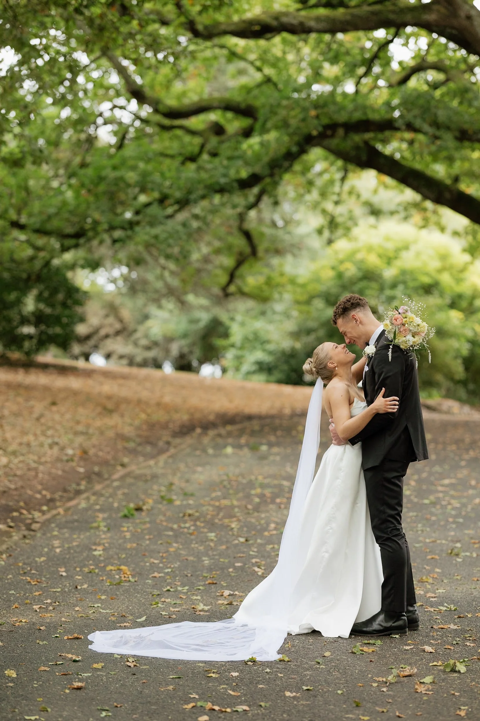 A bride and groom standing close together outdoors in a park, smiling and gazing into each other's eyes. The bride is in a white wedding gown with a long train, holding a bouquet of flowers. The groom is in a black suit and tie. They are surrounded by green trees and fallen leaves on the ground.