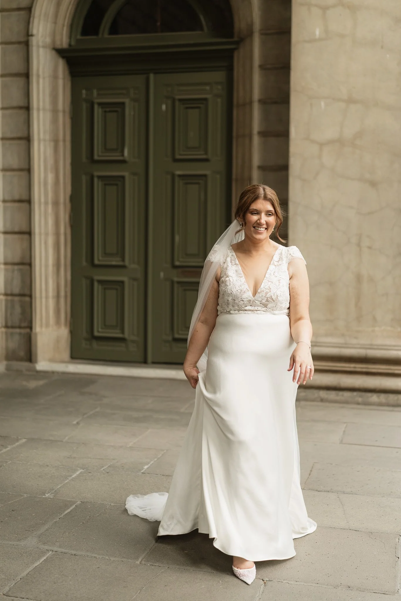 A smiling bride in a white wedding gown standing outdoors in front of a large green door and stone columns.