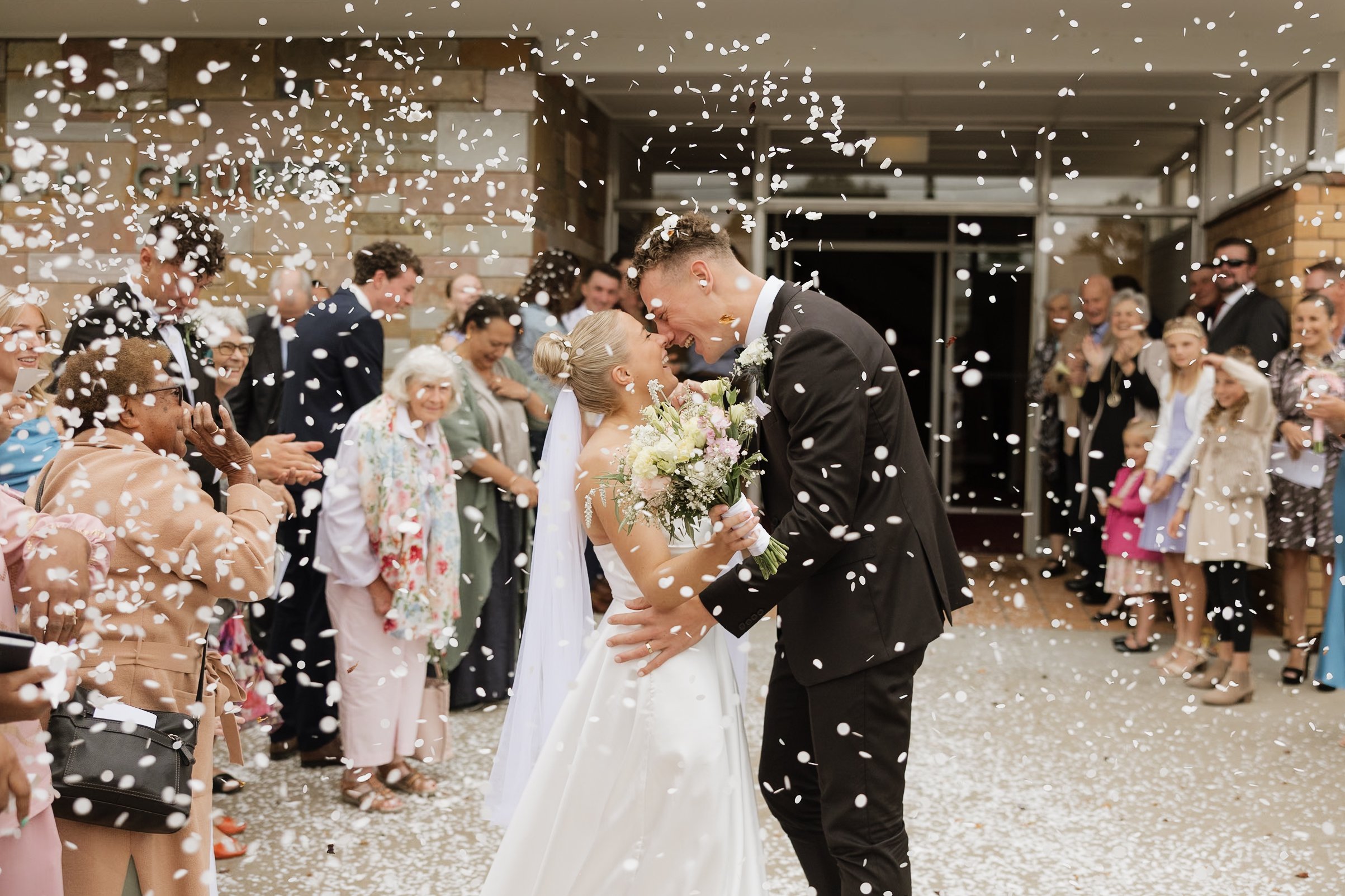 A newlywed couple shares a kiss under falling confetti during their wedding celebration, surrounded by smiling guests outside a venue.
