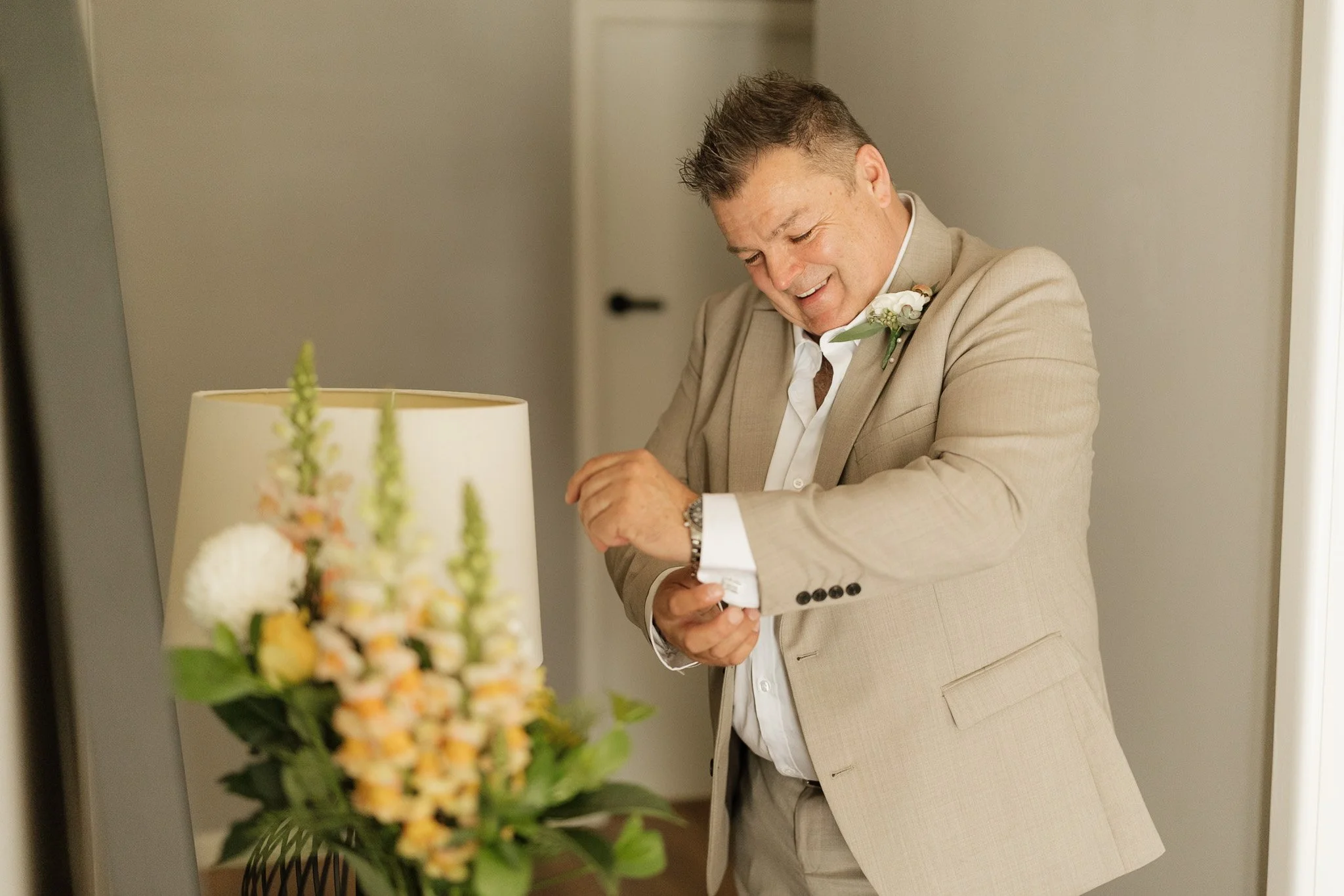 A man in a beige suit smiling and looking down, with a boutonniere, standing indoors near a door, with a blurred bouquet of flowers in the foreground.