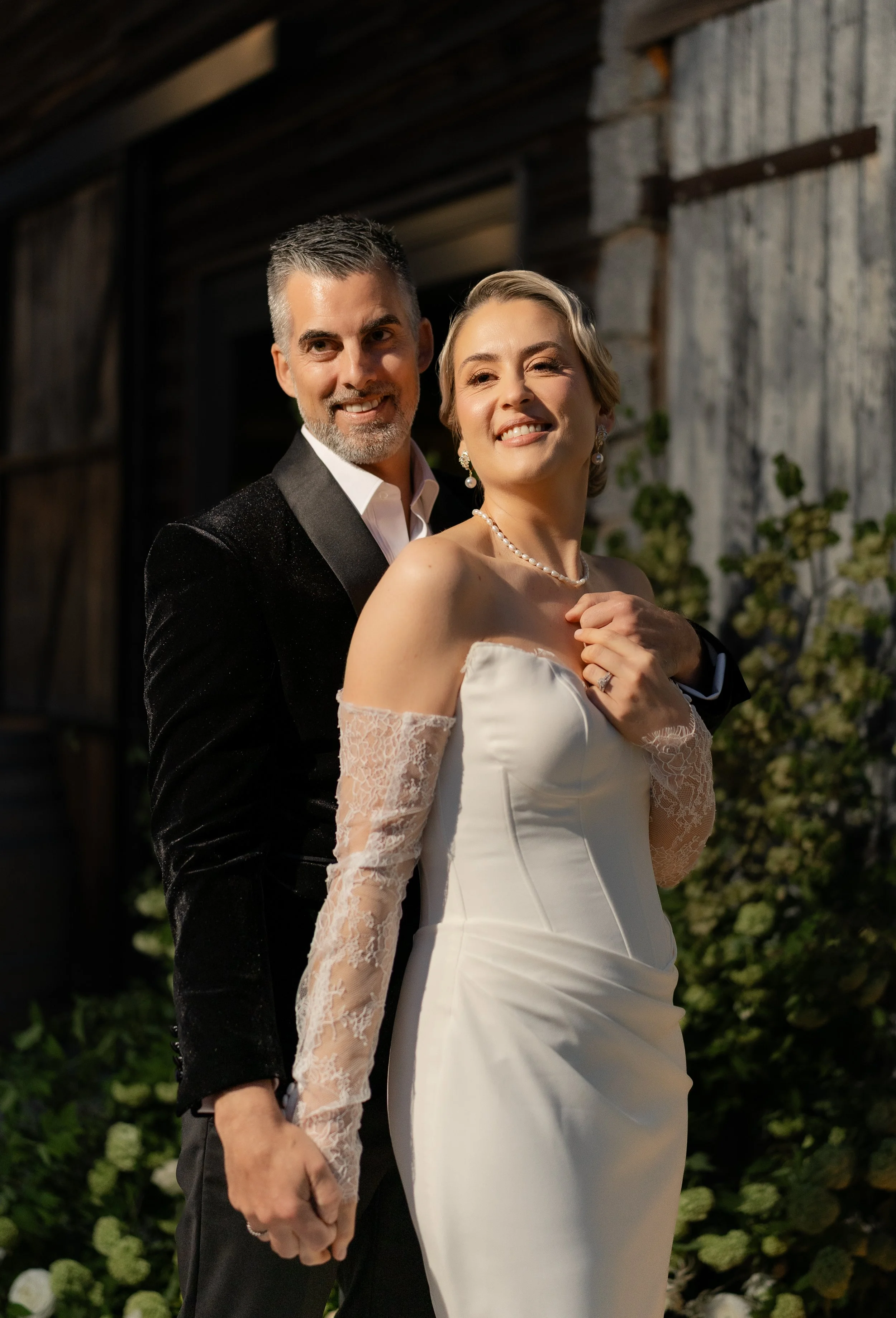 A newlywed couple dressed in wedding attire, standing outdoors, smiling at the camera, with greenery and rustic building background.
