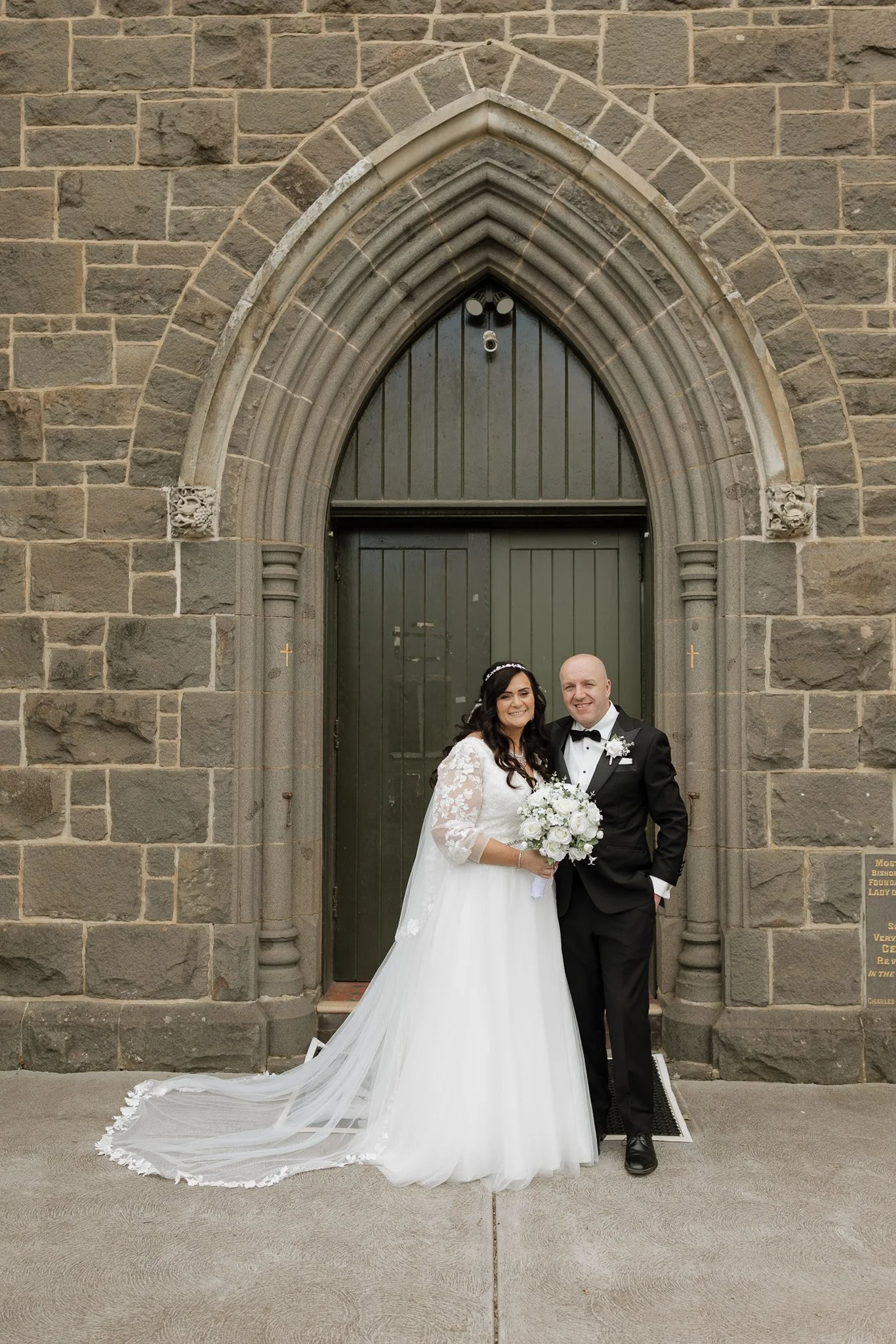 A newlywed couple standing in front of a stone church entrance, smiling and posing for a wedding photo. The bride is in a white wedding dress holding a bouquet, and the groom is in a black tuxedo with a bow tie.