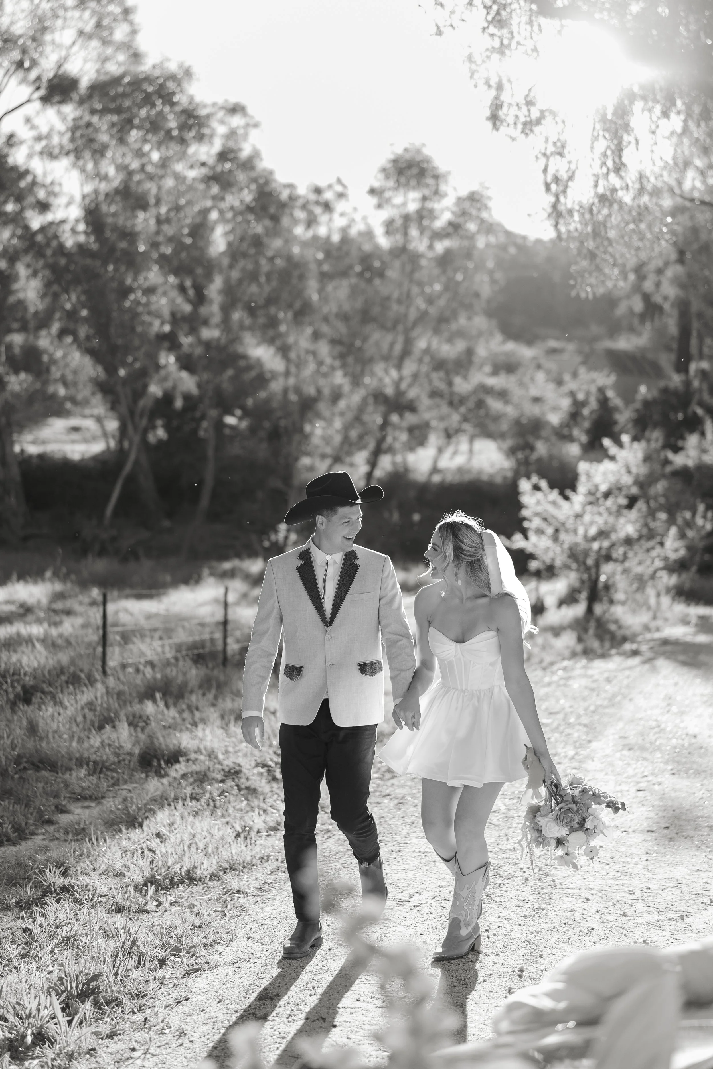 Black and white photo of a happy couple walking hand in hand outdoors, with the woman holding a bouquet of flowers and wearing cowboy boots and a strapless dress, while the man is in a suit jacket and cowboy hat, surrounded by trees and sunlight.