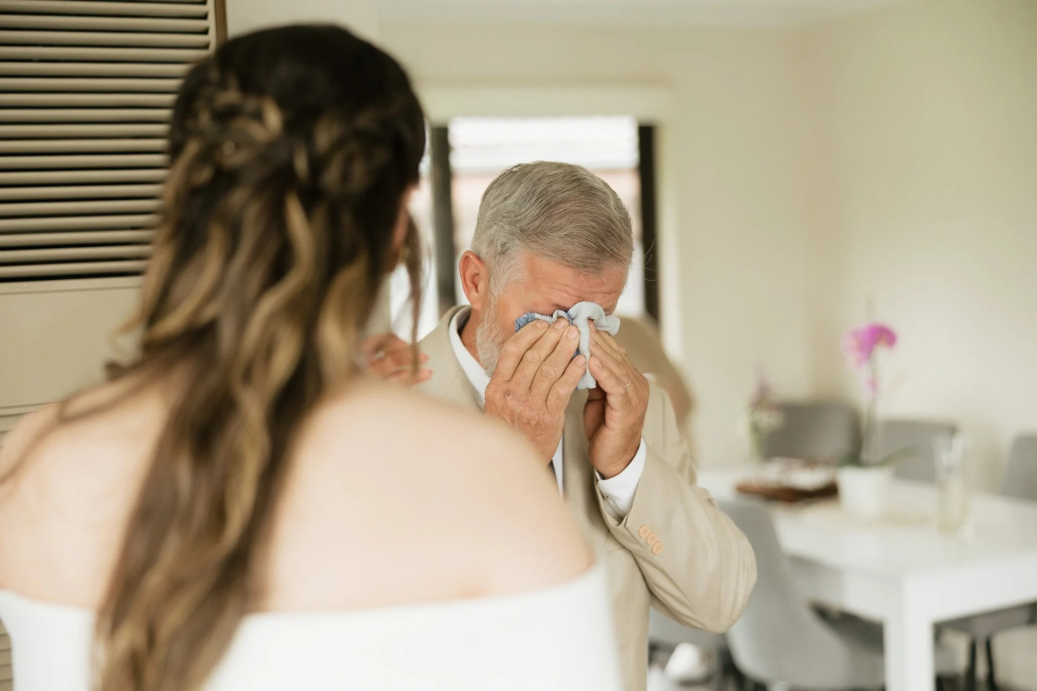 A man with gray hair and a beige suit is crying and wiping his eyes with a tissue. A woman with long, wavy brown hair is in the foreground, facing away from the camera.