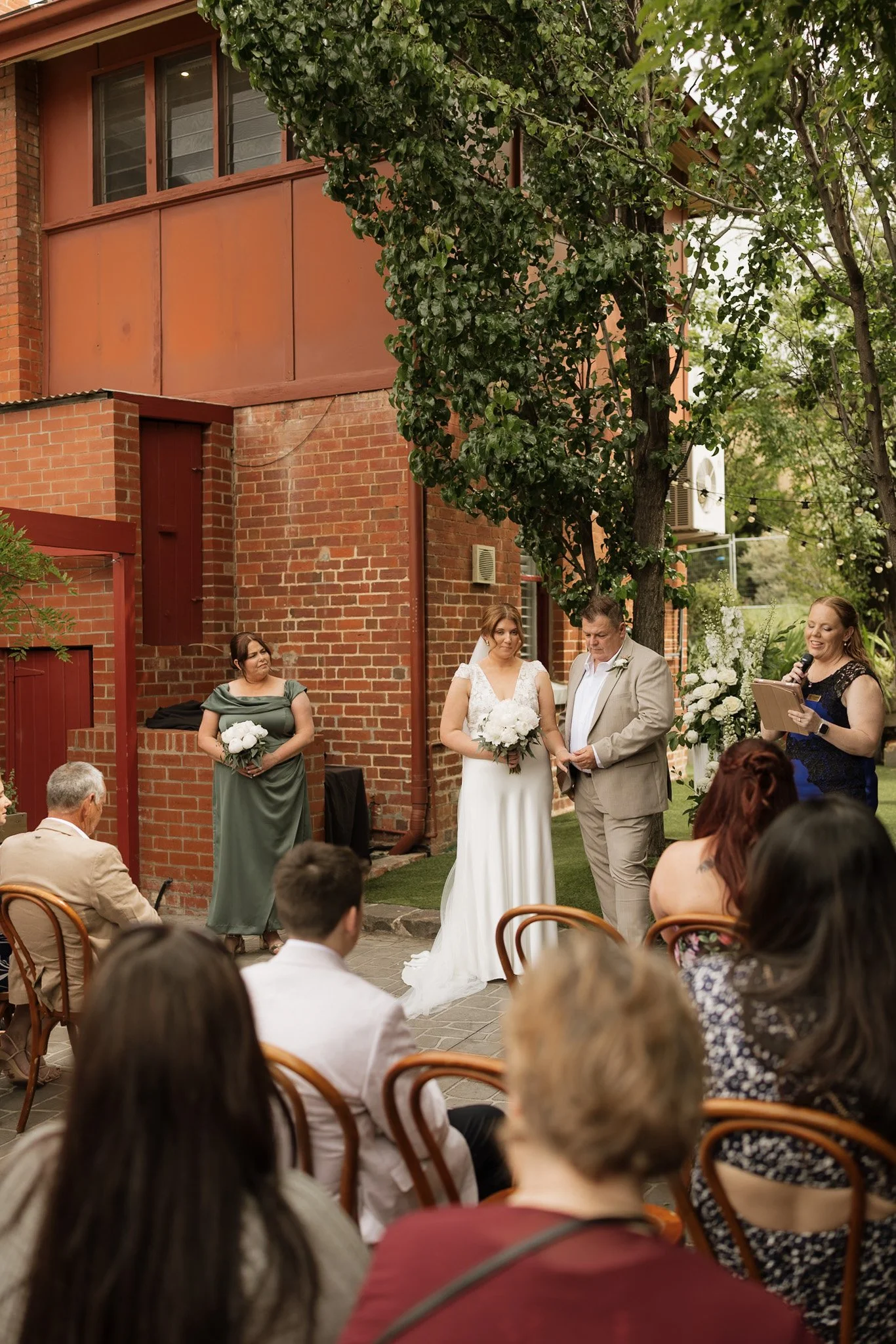 A wedding ceremony taking place outdoors on a brick patio with a brick building and green trees in the background. The bride and groom are standing together, the bride in a white dress and holding a bouquet, and the groom in a light suit. A woman in 