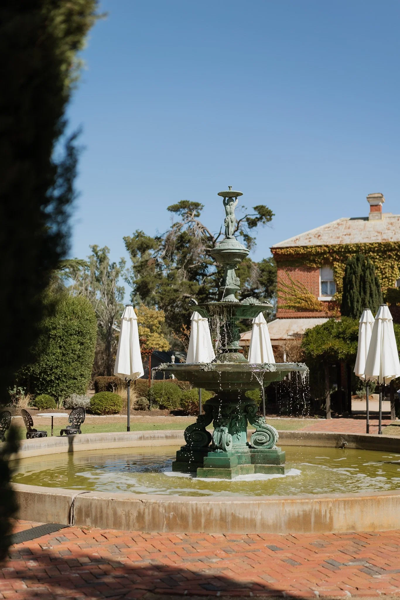 A green ornate fountain with a statue of a woman holding a shell on top, water flowing from it, surrounded by white umbrellas and garden benches, with trees and a brick building in the background.