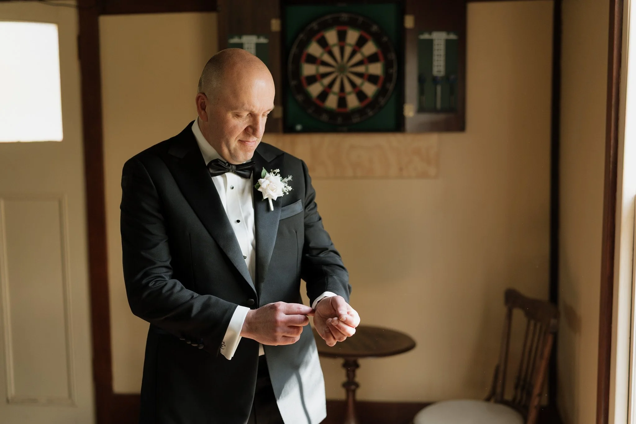 A man dressed in a tuxedo with a white flower boutonniere is looking down as he adjusts his cuffs in a room with beige walls. In the background, there is an electronic dartboard on the wall and a small wooden table.