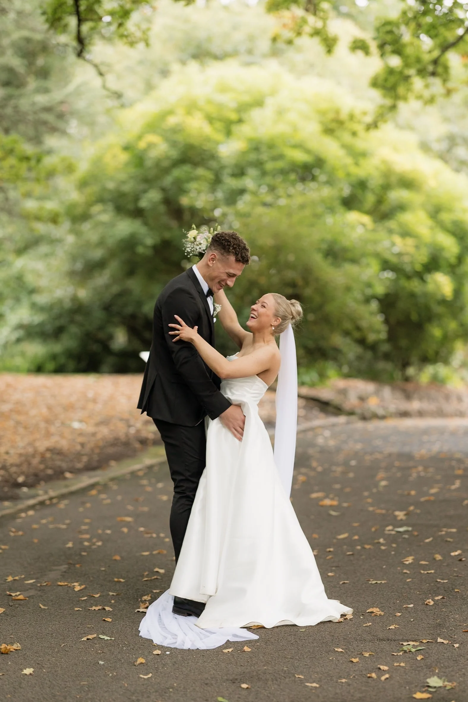 A newlywed couple dancing outdoors on a paved path covered with fallen autumn leaves, surrounded by green trees. The groom, dressed in a black tuxedo, and the bride, in a strapless white wedding dress with a long veil, are smiling and sharing a joyfu