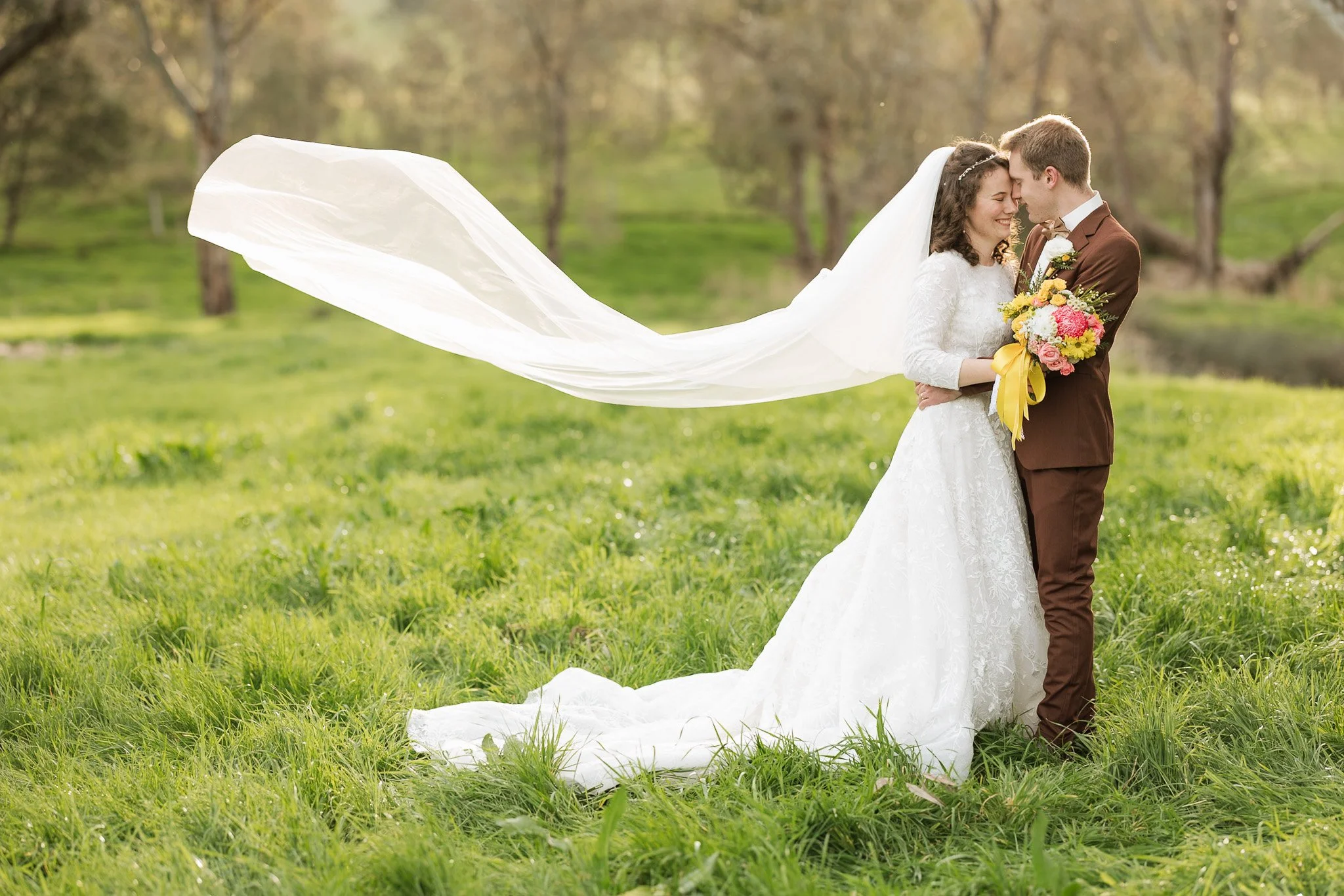 A bride and groom in wedding attire standing close together outdoors, touching foreheads and smiling.