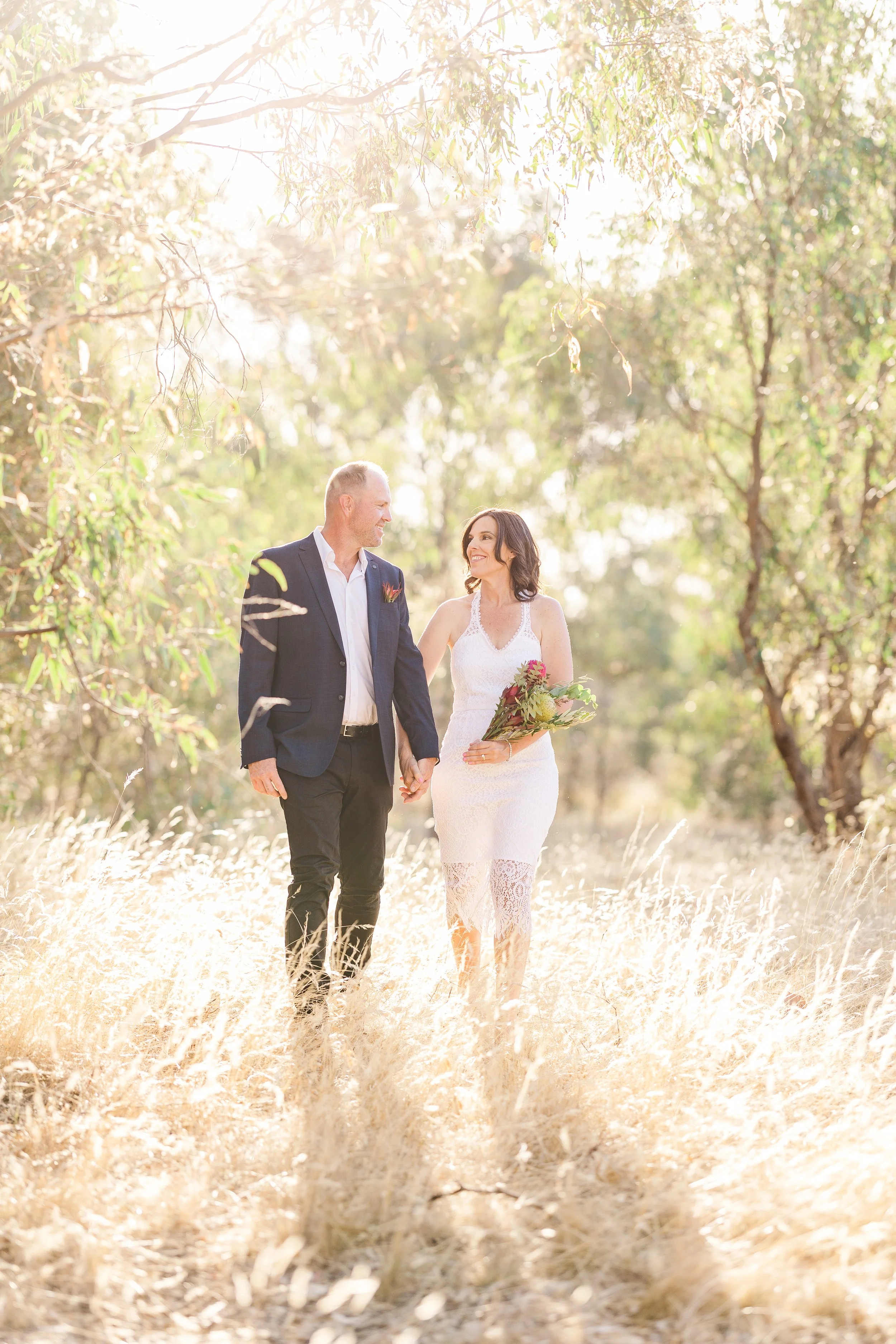 A happy couple walking hand in hand through a sunlit field, with the woman holding a bouquet of flowers, in a natural outdoor setting.