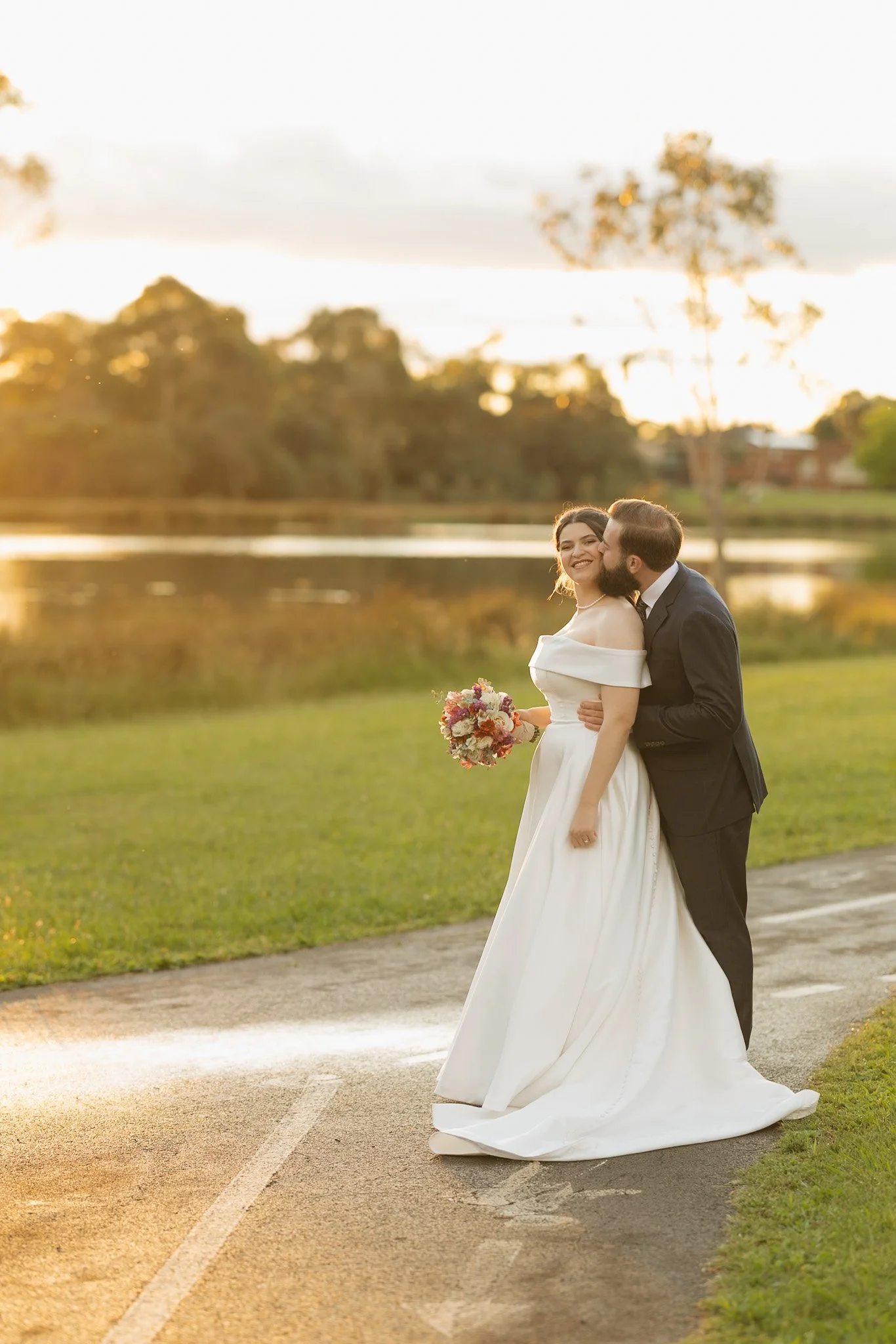 A newlywed couple dressed in wedding attire, embracing outside during sunset. The bride is holding a bouquet of flowers and smiling, while the groom kisses her cheek. The background features trees and a body of water.