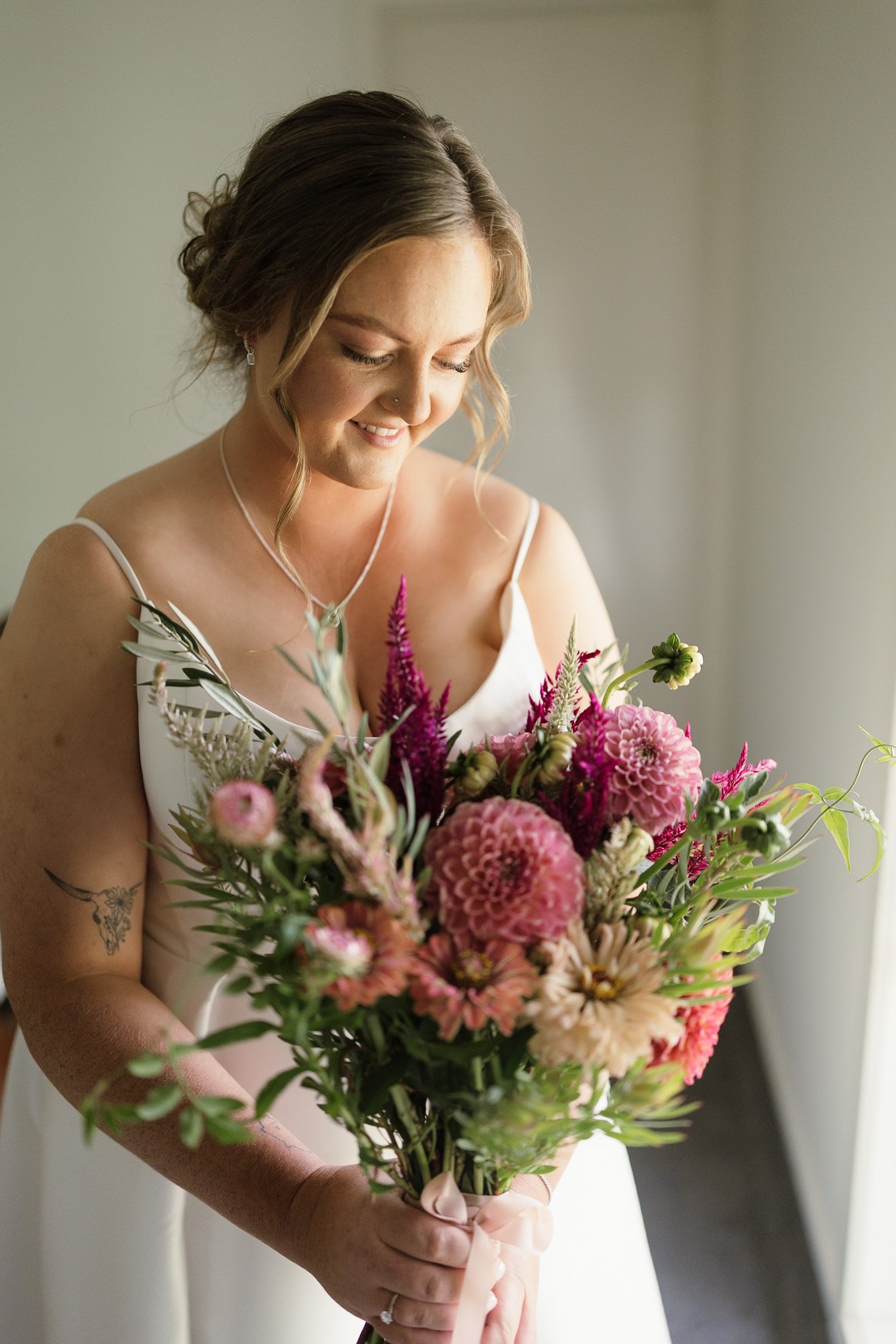 A woman in a white dress holding a large bouquet of pink, purple, and white flowers, smiling with her eyes closed.