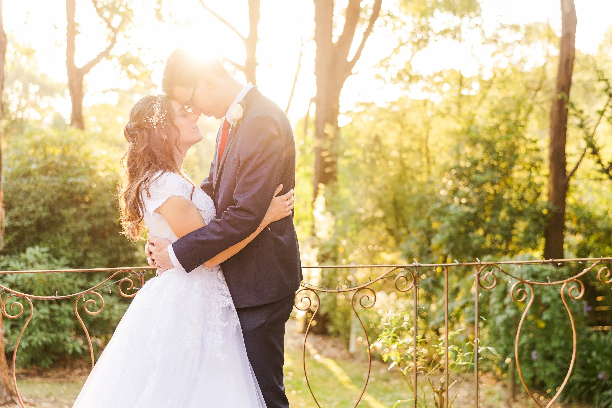 A bride and groom sharing a romantic moment outdoors during sunset, with the bride in a white wedding dress and the groom in a suit, surrounded by trees and a decorative fence.