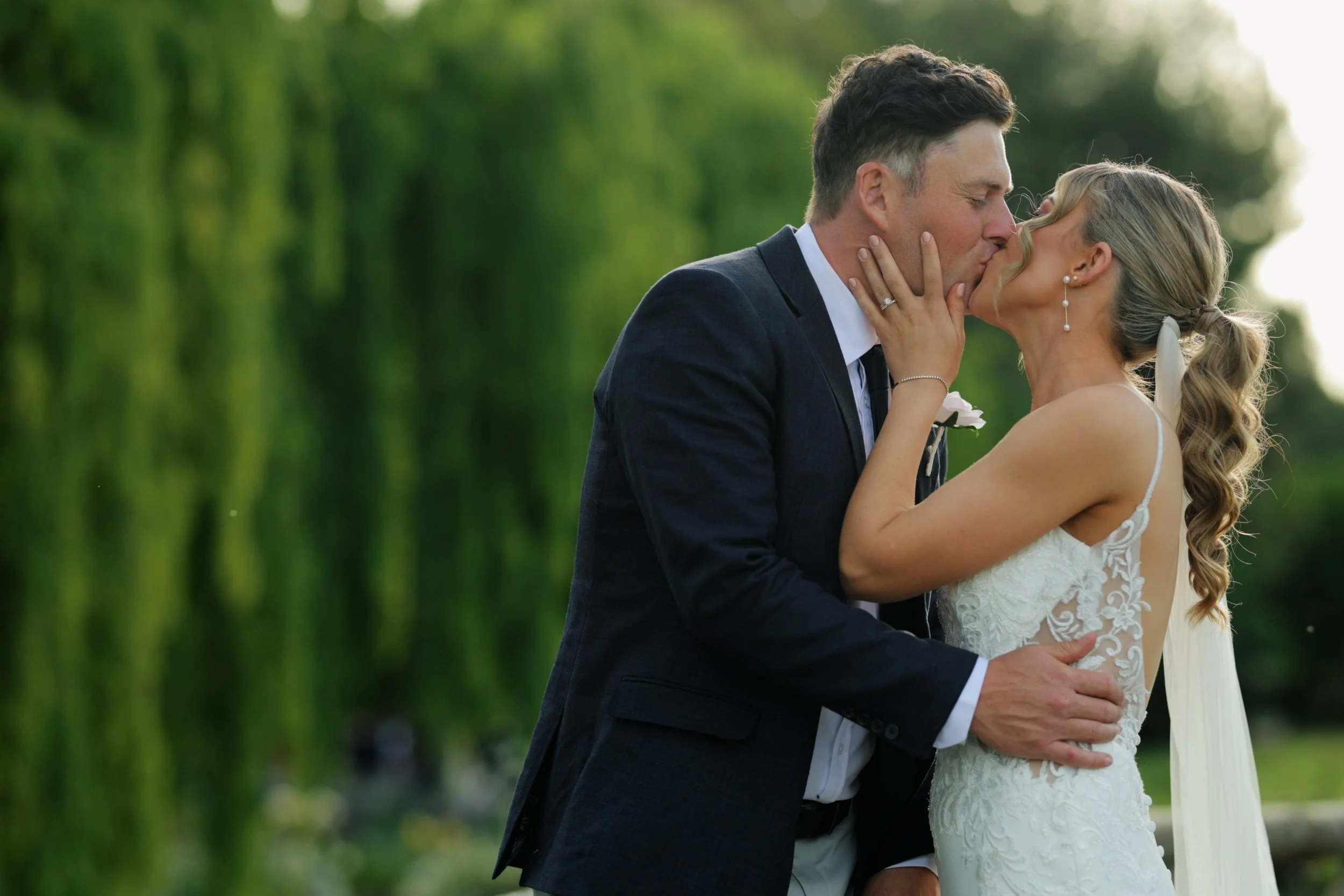 A bride and groom share a kiss outdoors during their wedding, with greenery in the background.