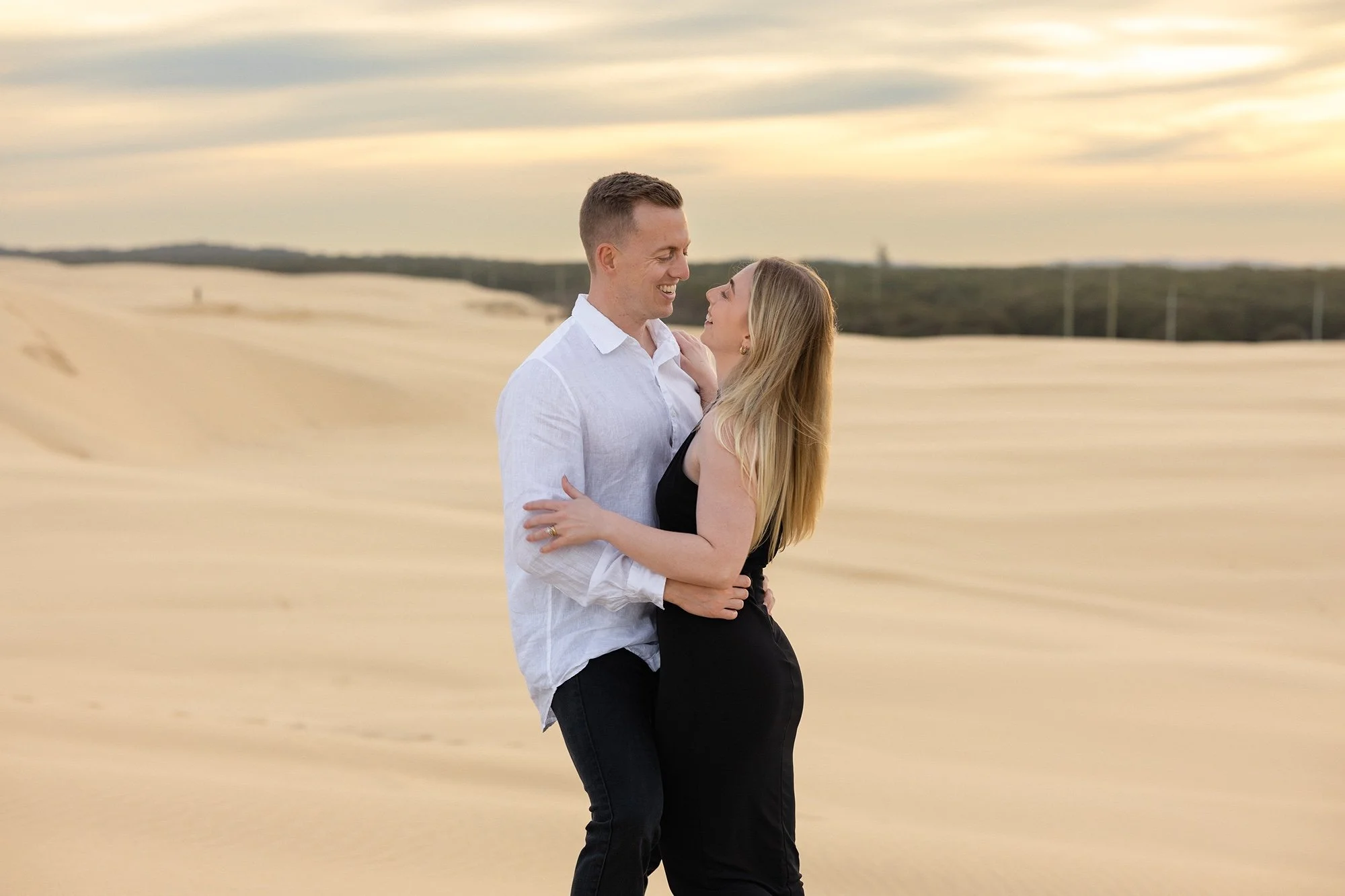 A couple stands close and embraces in a sandy desert landscape at sunset, looking into each other's eyes and smiling.